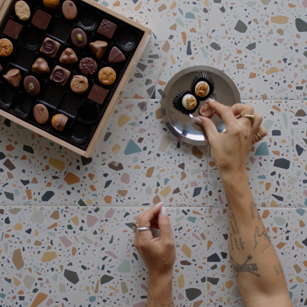 Hands placing chocolates on a small plate over terrazzo-style large format tiles with multicolored fragments