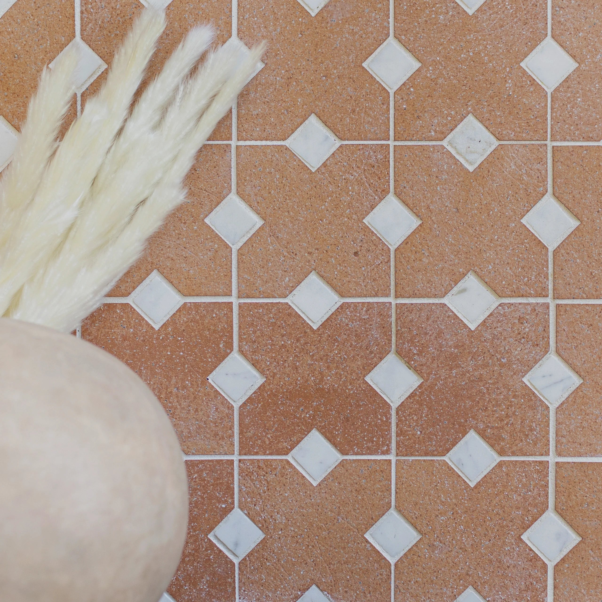 Pattern of terracotta tiles with white diamond-shaped accents next to neutral-toned dried pampas grass in round vase