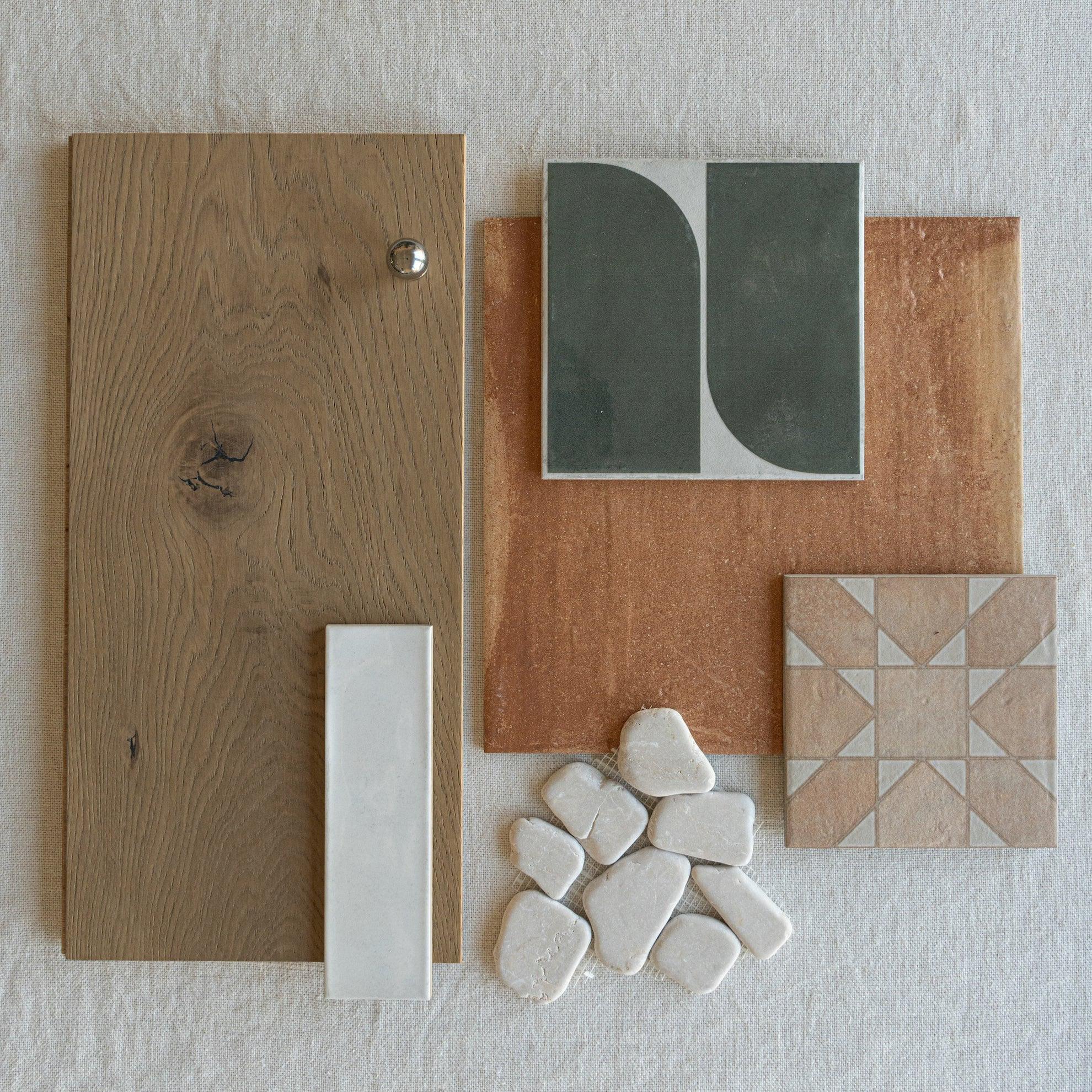 Flat lay featuring oak wood panel with knob, terracotta tile, geometric ceramic tiles, and smooth white stone samples on fabric background