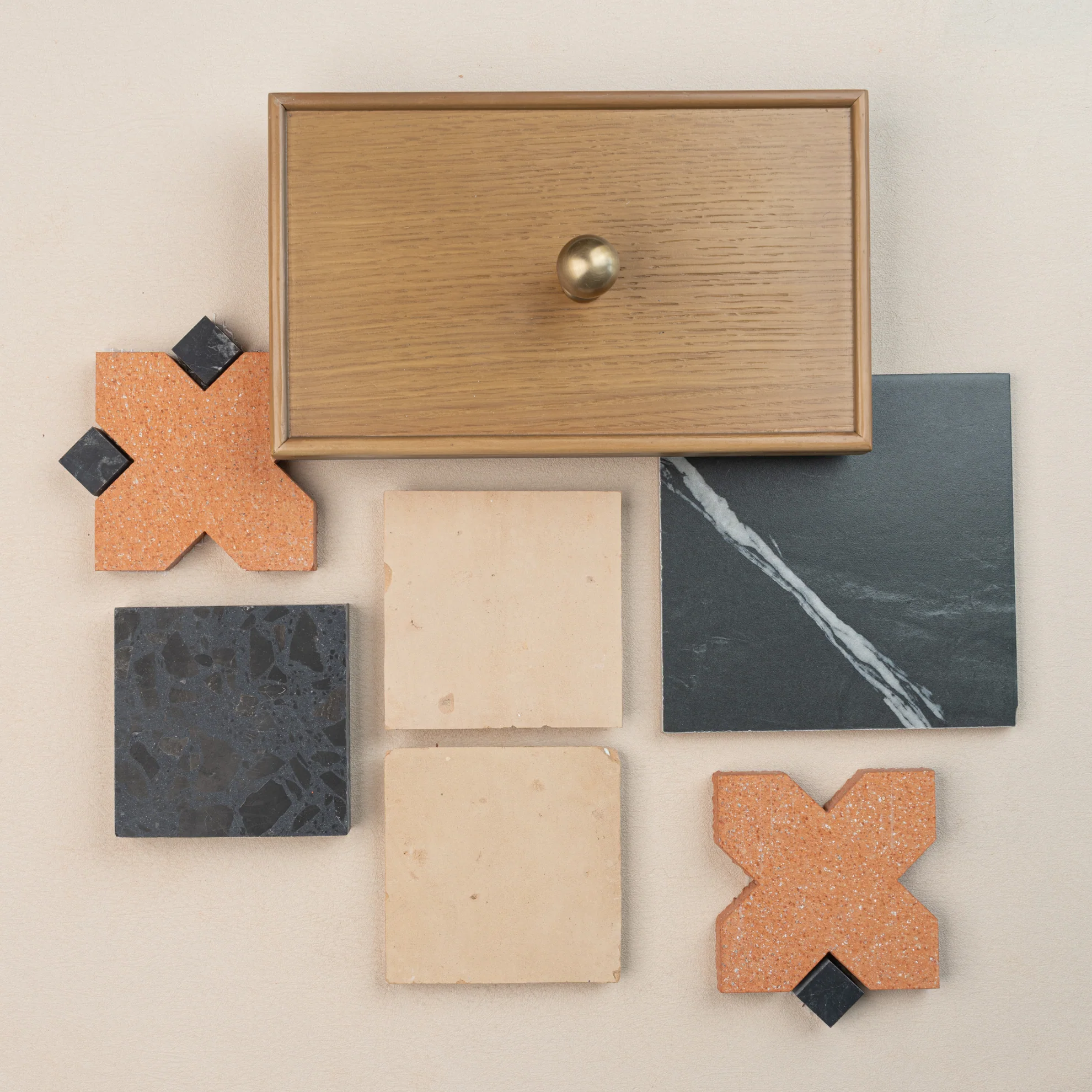 Flat lay of oak wood cabinet drawer front with brass knob, black marble tile, beige stone tiles, and terracotta ceramics on pale surface