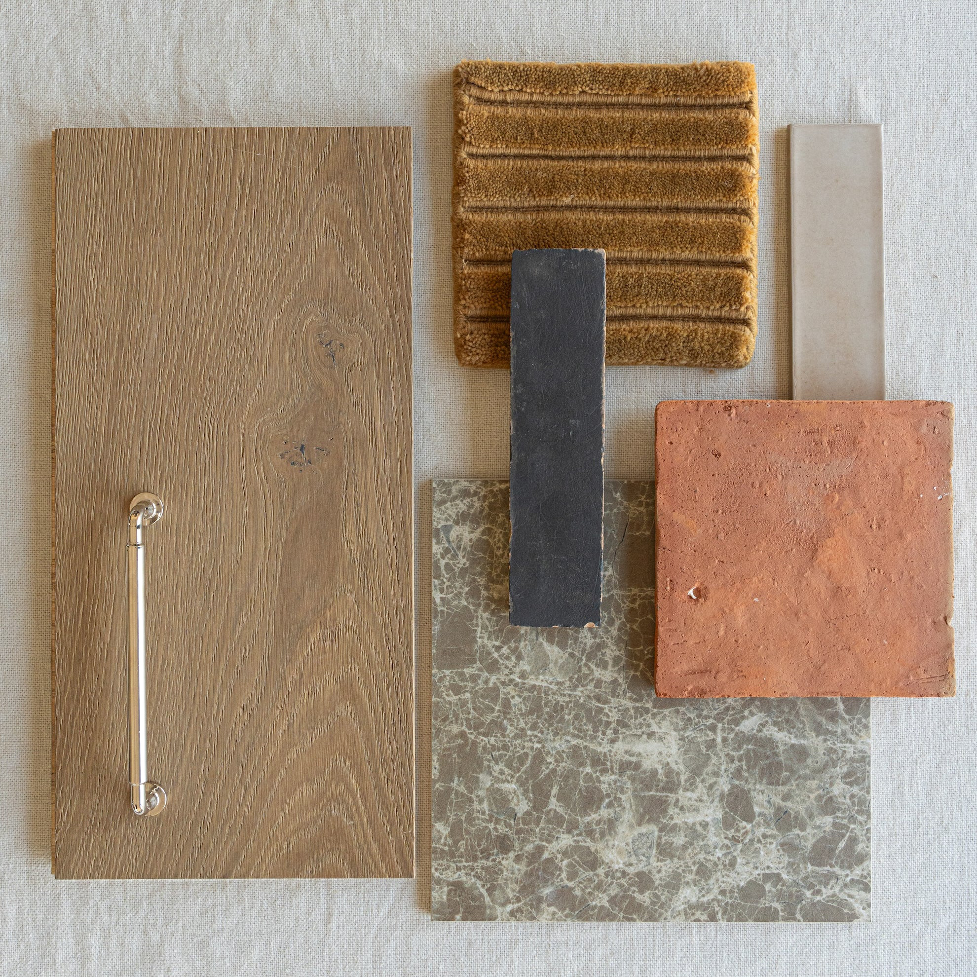 Flat lay of oak wood cabinet door with metal handle, terracotta tile, textured fabric, dark rectangular tile, and marbled tile sample on neutral background