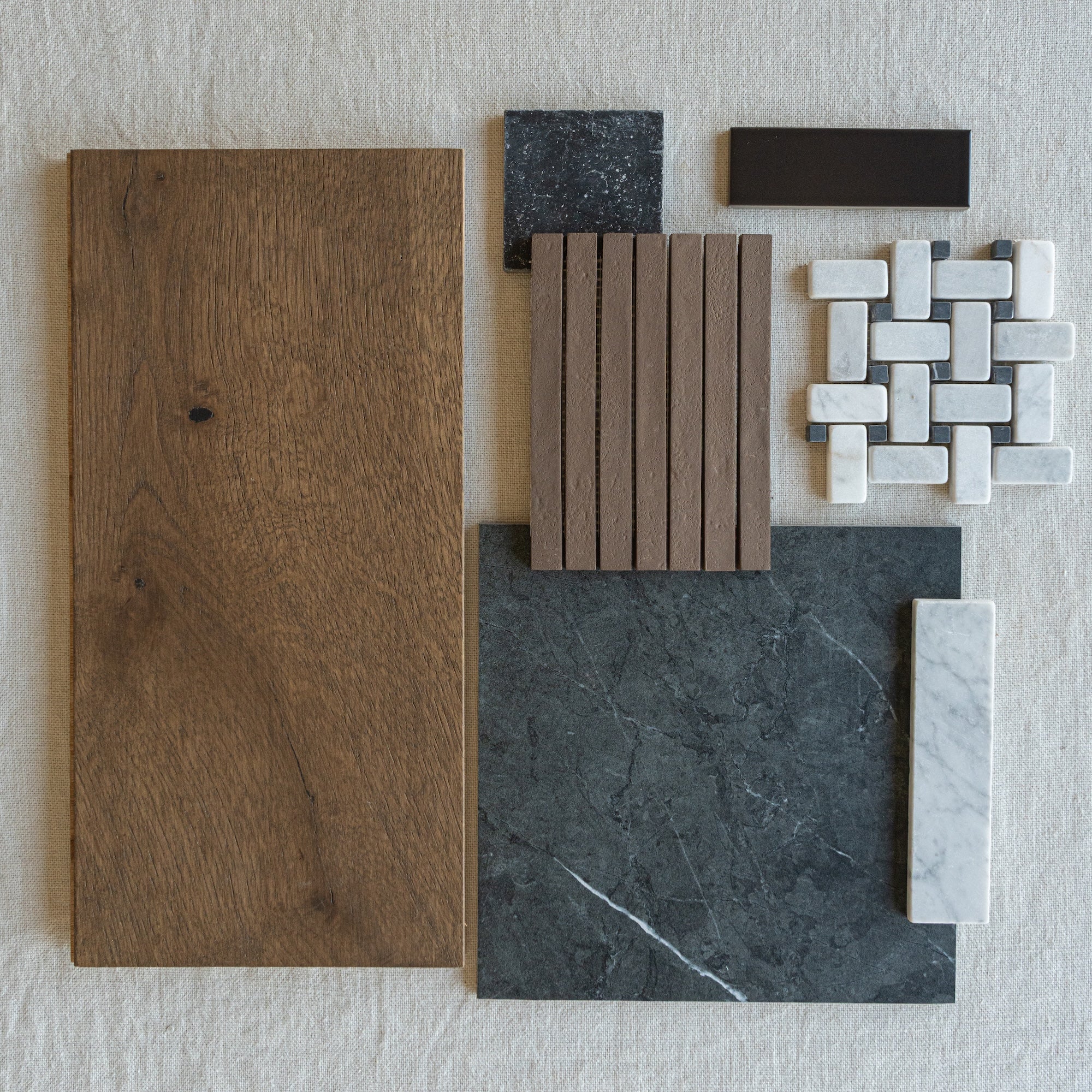 Flat arrangement of oak wood plank, dark stone tile, white marble mosaic, brown ridged slats, and dark rectangular tile on fabric background