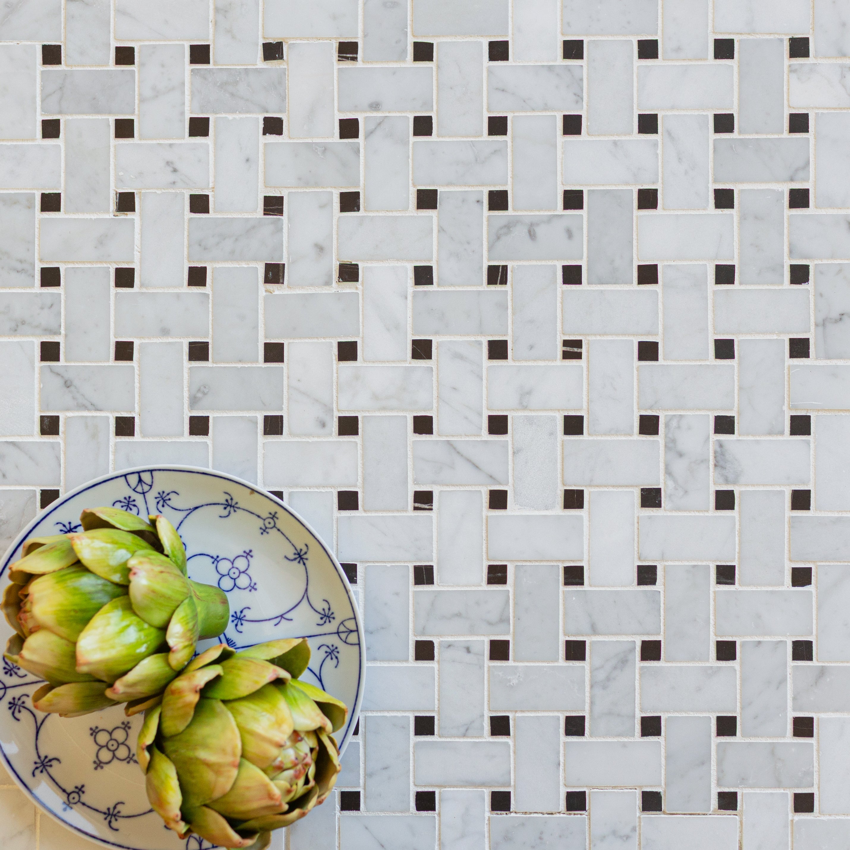 White marble basketweave tiles with small dark square accents and a blue patterned plate holding two artichokes