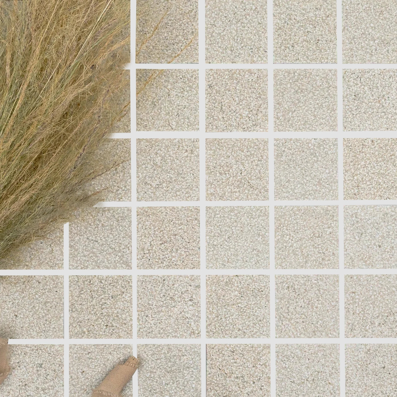 Grid of small square tiles with a sandy beige texture arranged beside dried ornamental grasses in a neutral setting