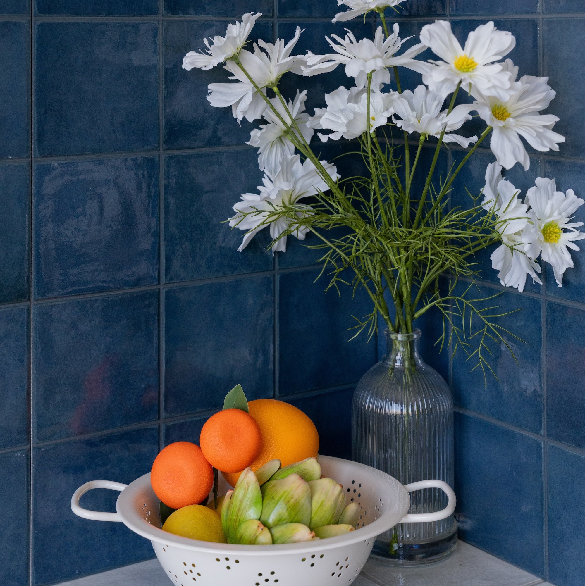 White daisies in a glass vase beside a colander of fresh citrus and vegetables against blue tiled walls