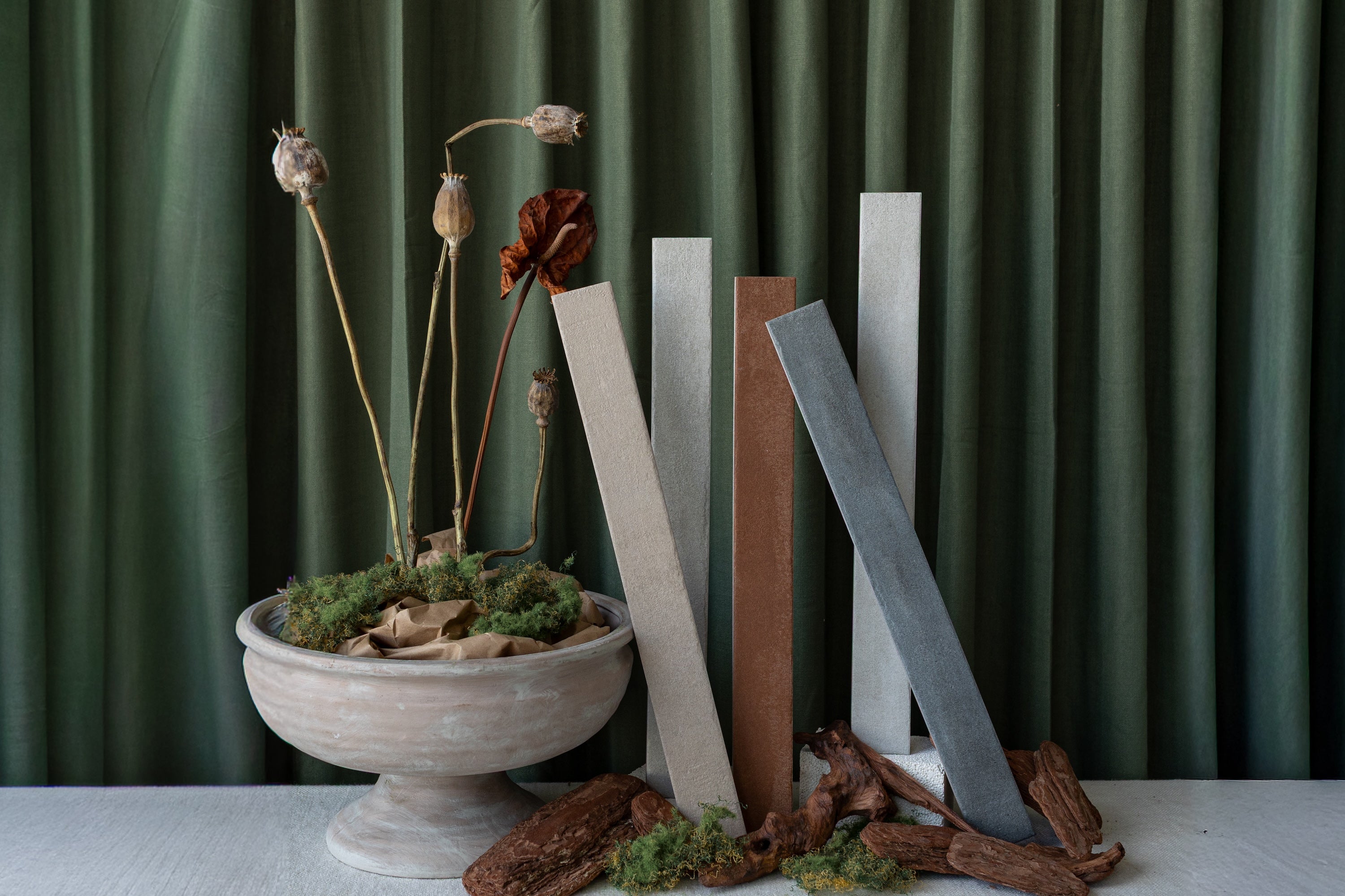 Rectangular tile samples in muted beige, brown, and gray tones arranged with dried pods and moss in a stone bowl against a green curtain backdrop