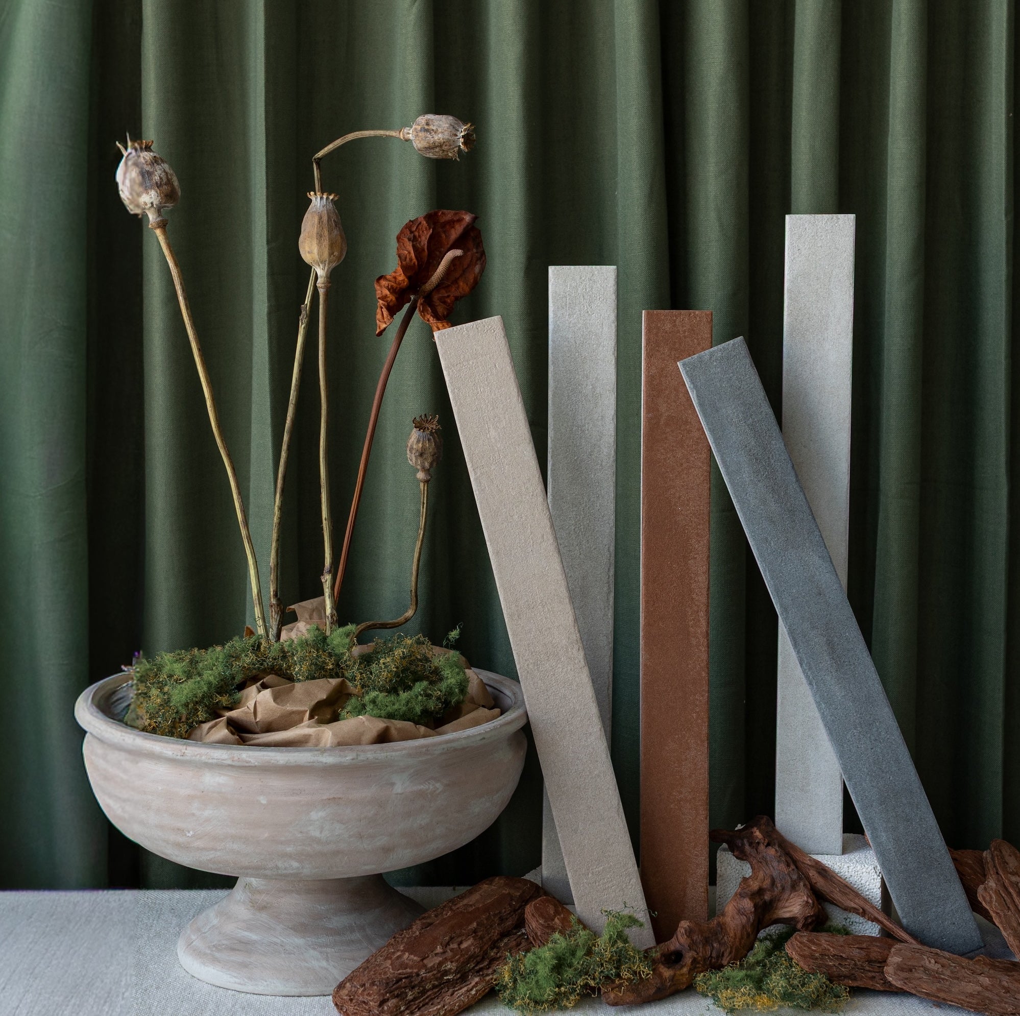 Tall neutral ceramic tile samples displayed upright next to dried floral stems in a rustic pot on a fabric surface