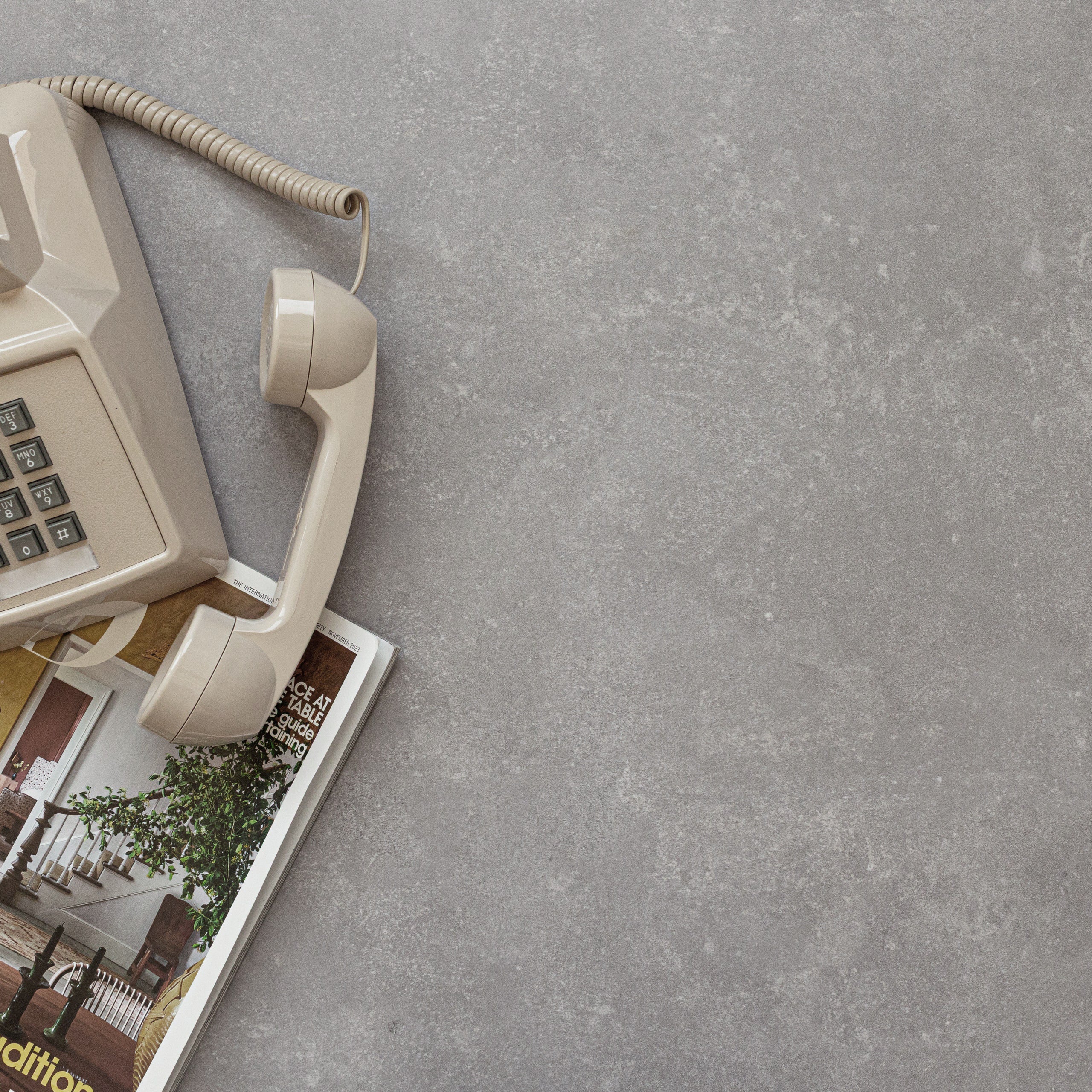 Vintage beige corded telephone handset resting on a gray cement-look porcelain tile surface with an interior design magazine below.