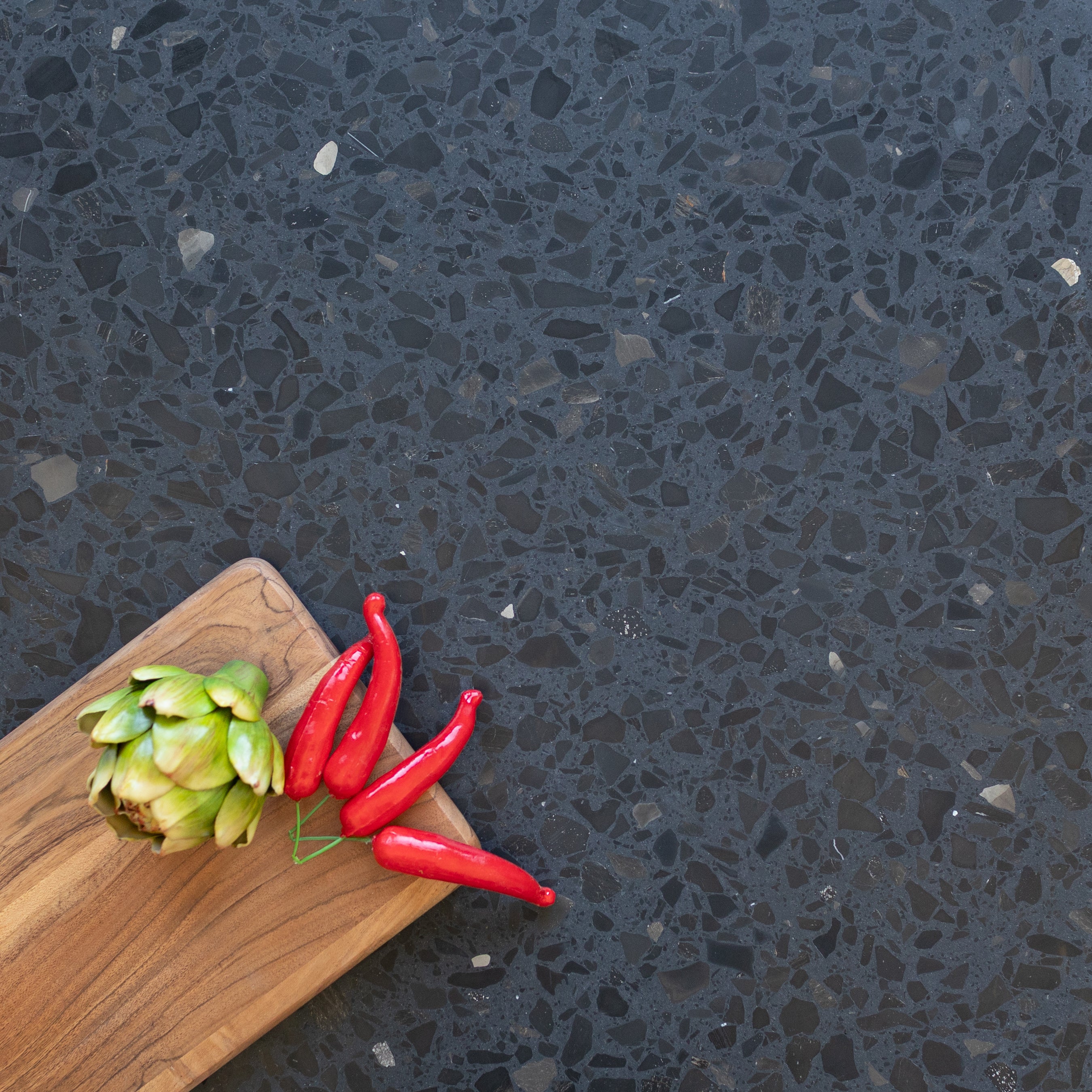 Dark terrazzo surface with mixed black and gray stone fragments, featuring a wooden board with red chili peppers and an artichoke