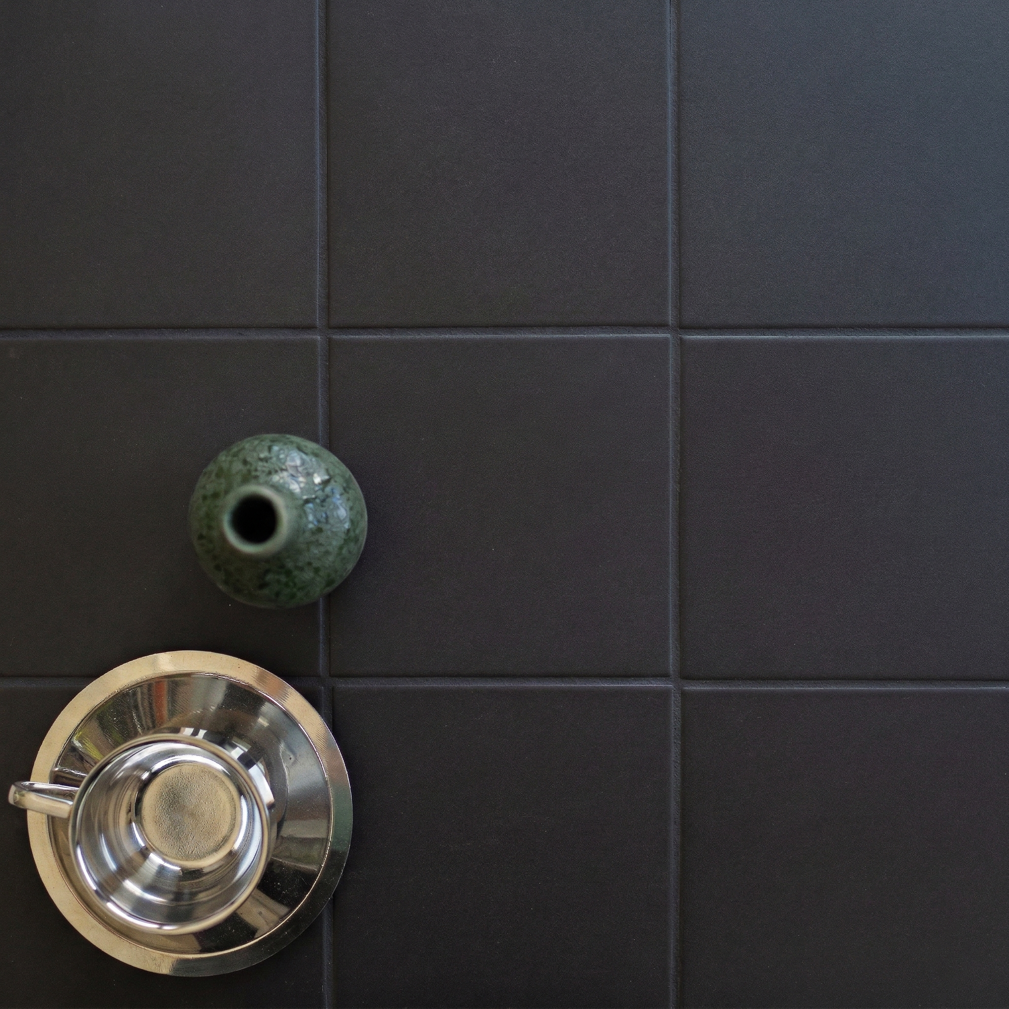 Minimalist setting with matte black ceramic tiles, a reflective metal cup, and a small green ceramic vase on a dark surface