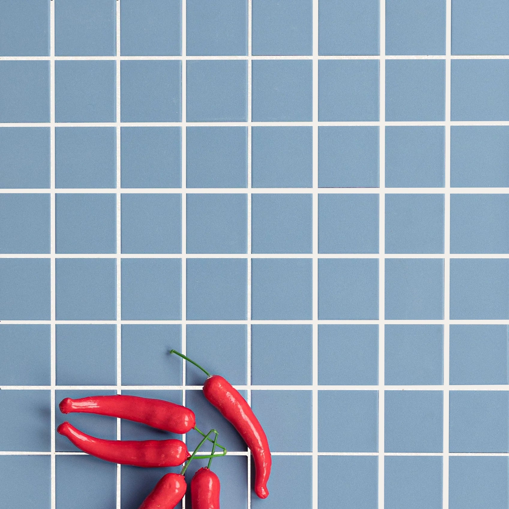 Matte blue small square porcelain tiles with white grout lines and several red chili peppers resting on the surface