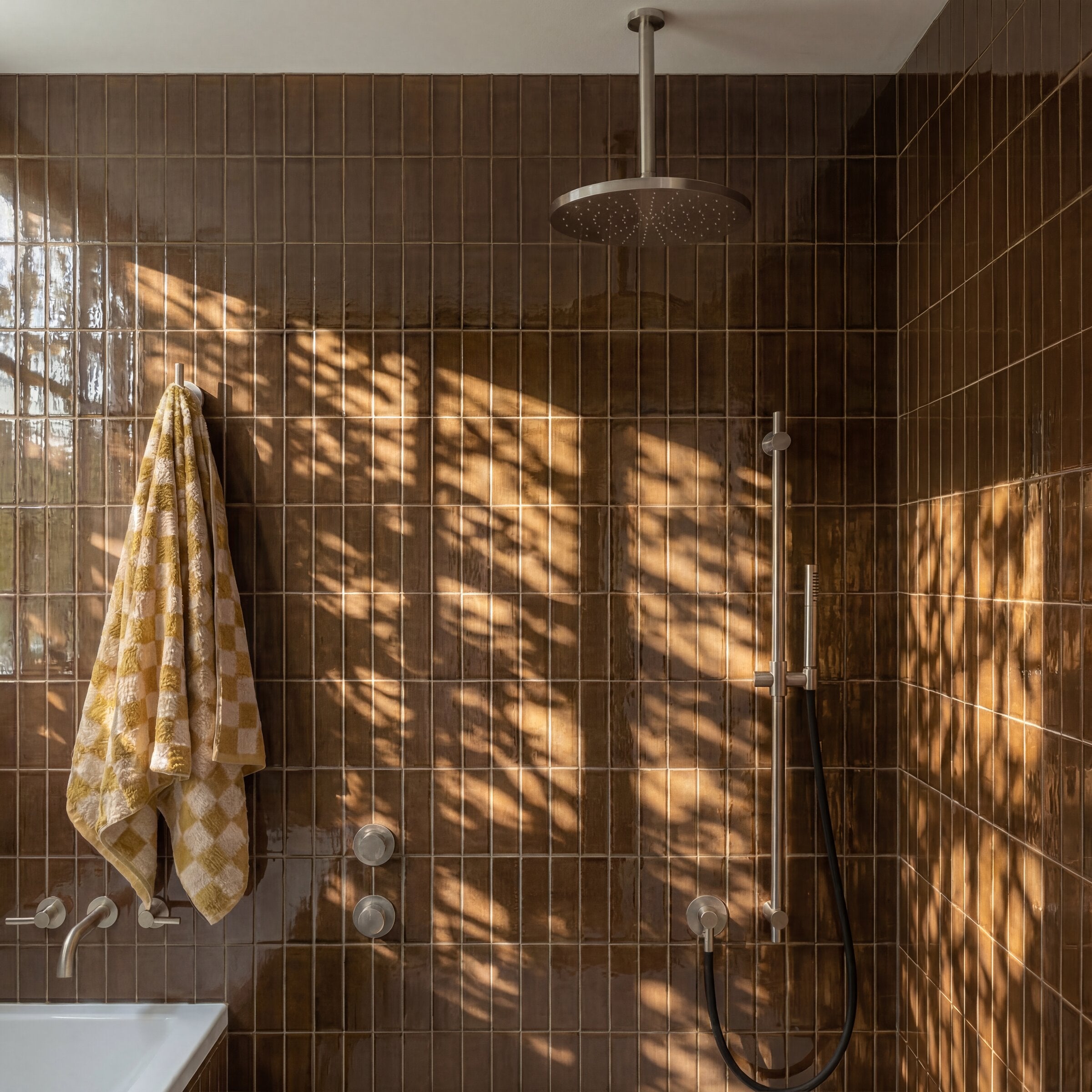 Glossy deep brown ceramic tile shower with overhead rain showerhead, wall-mounted controls, and patterned towel hanging nearby