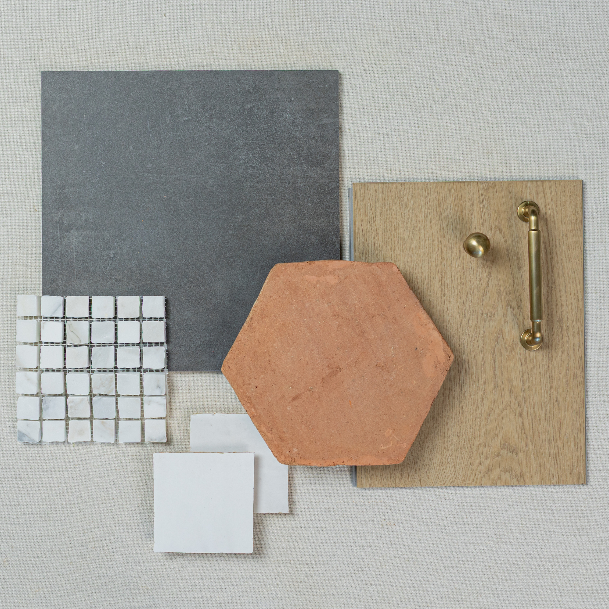Flat lay of matte grey stone-look tile, terracotta hexagonal tile, white ceramic mosaic, small white tiles, and light oak wood with brass hardware