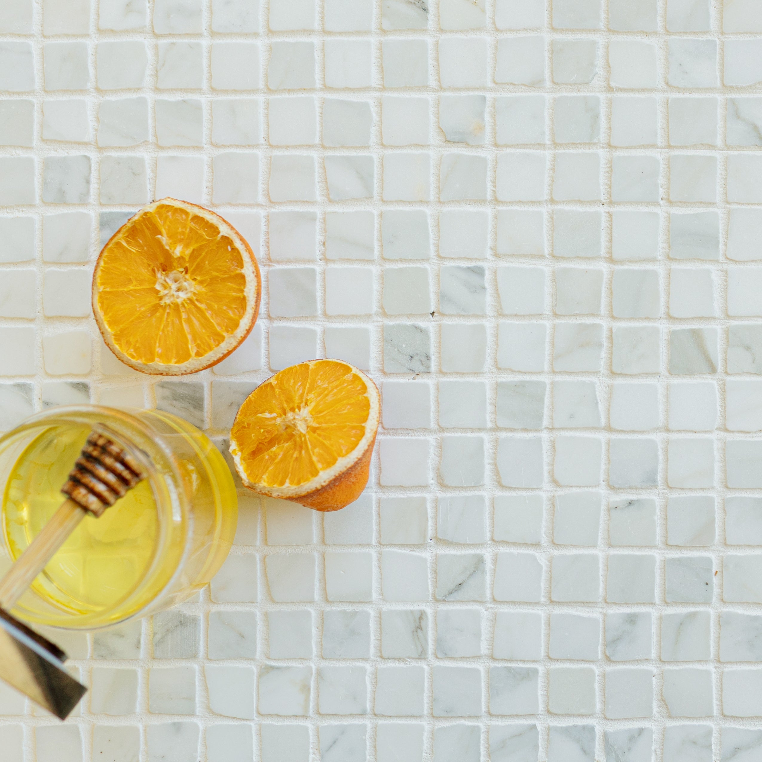 Small white marble mosaic tiles with subtle gray tones, a jar of honey with dipper, and two halves of an orange nearby