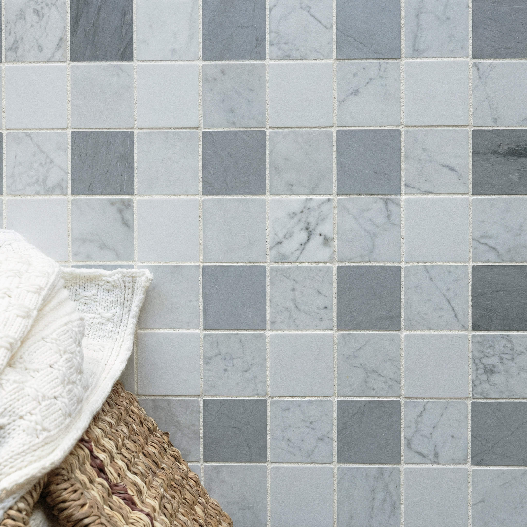 Square mosaic tiles in shades of gray and white with varied marble patterns beside woven basket and knitted blanket