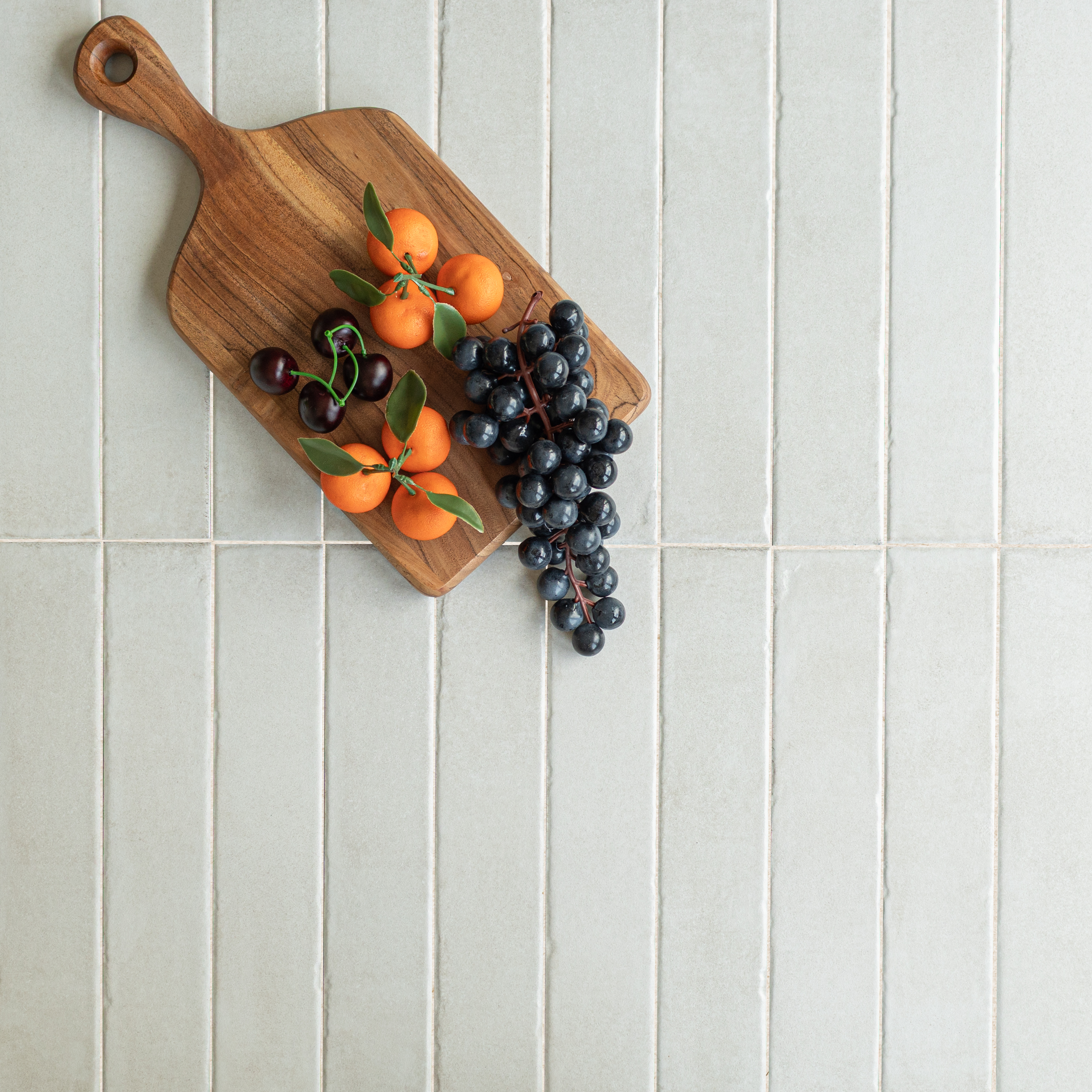 Top view of natural wood cutting board with clusters of cherries, tangerines, and black grapes on pale textured tiles