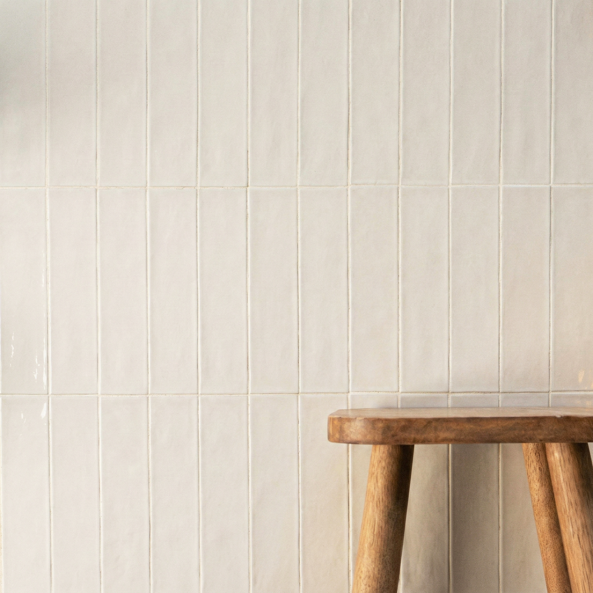 Cream-colored vertical ceramic tiles with a subtle glossy finish next to a rustic wooden stool leg on a neutral wall