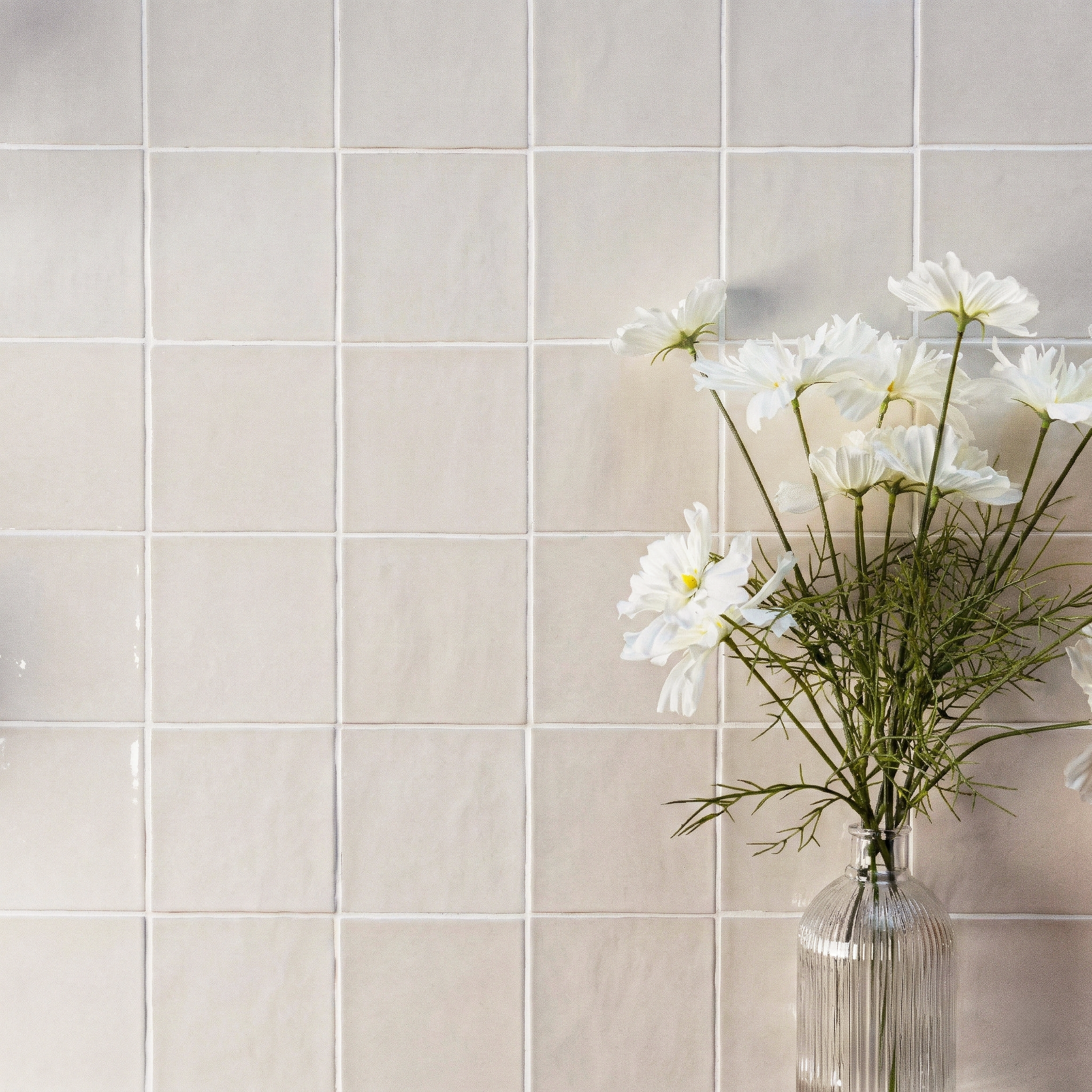 Cream-toned glazed ceramic tiles with a subtle handcrafted texture behind a clear ribbed glass vase holding white daisies