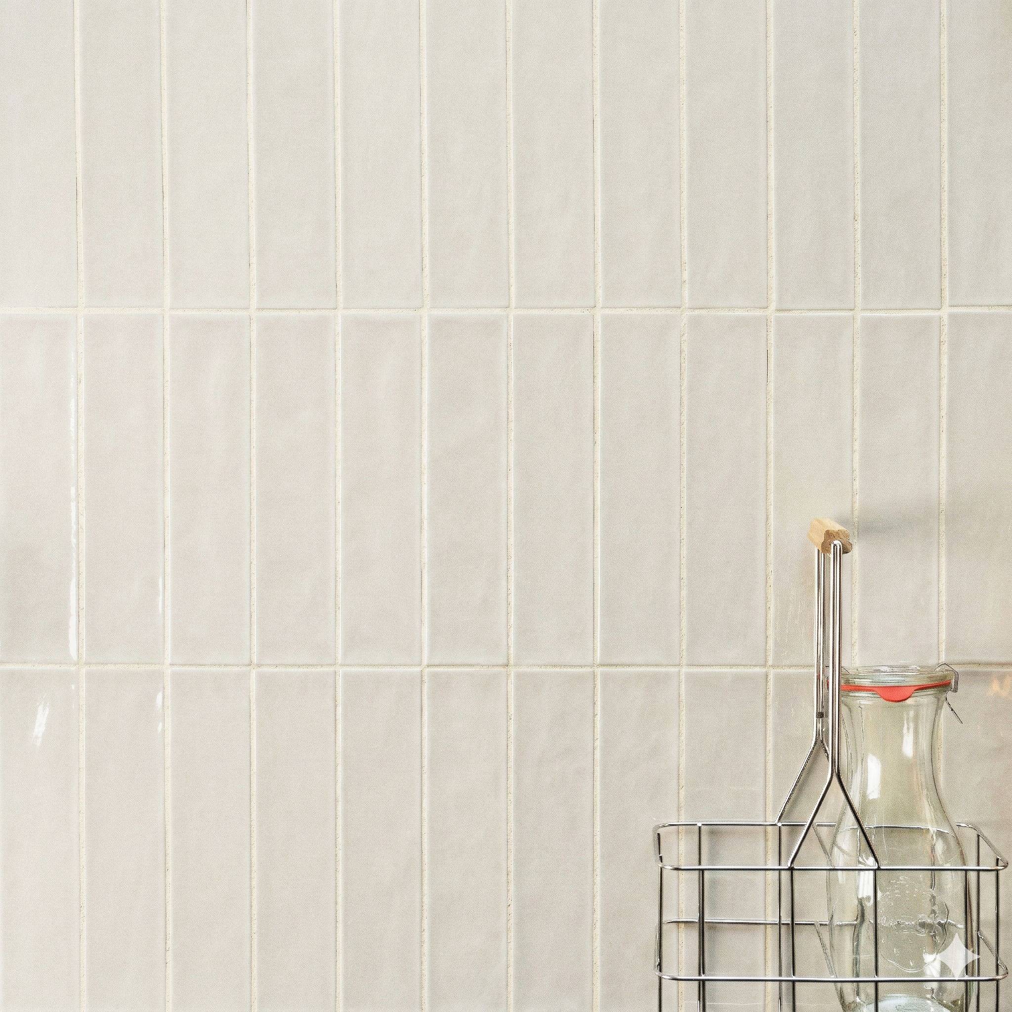 Glossy off-white rectangular ceramic tiles with subtle texture behind a glass bottle in a metal wire holder