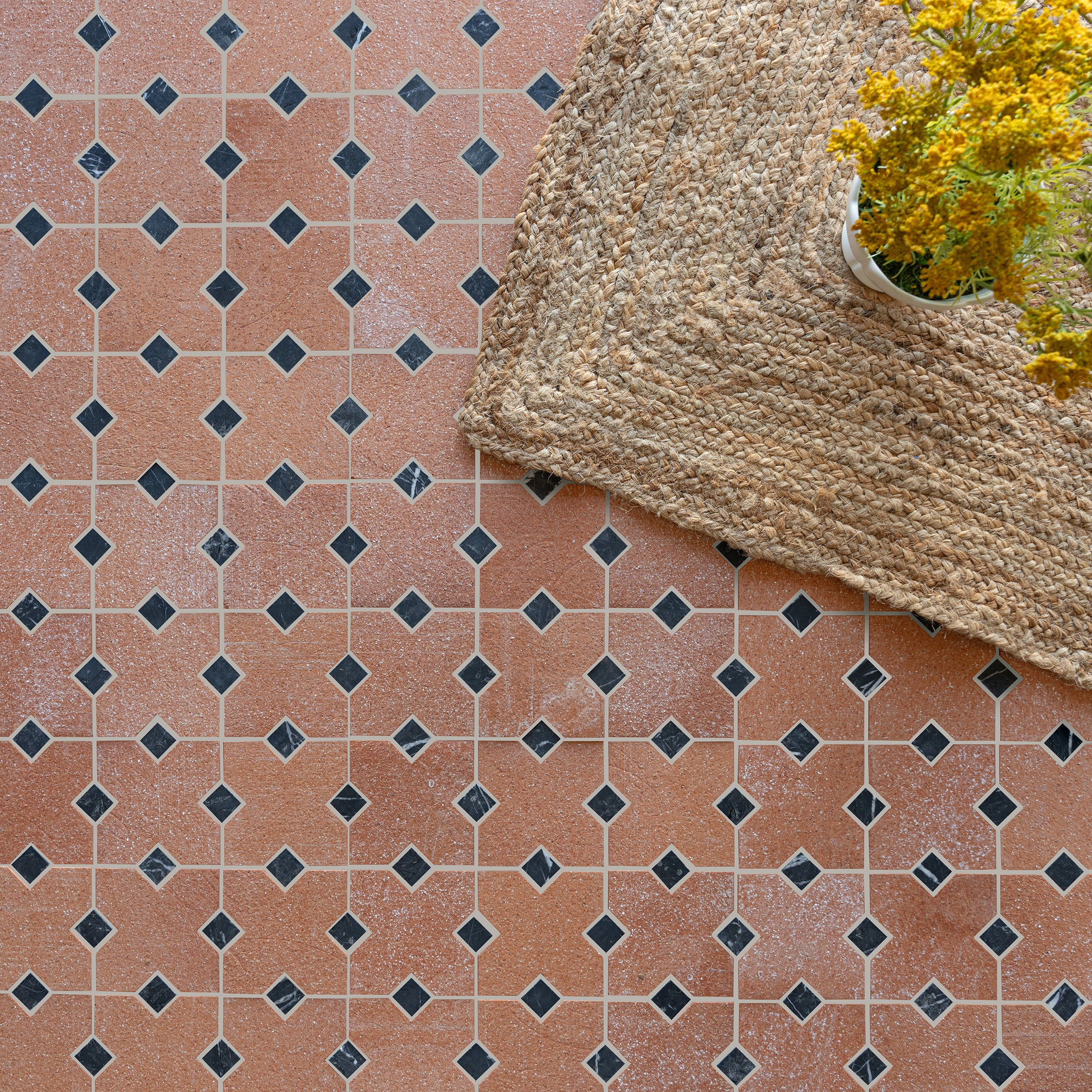 Terracotta ceramic tiles with black diamond accents and a natural woven rug partially covering the floor next to yellow flowers