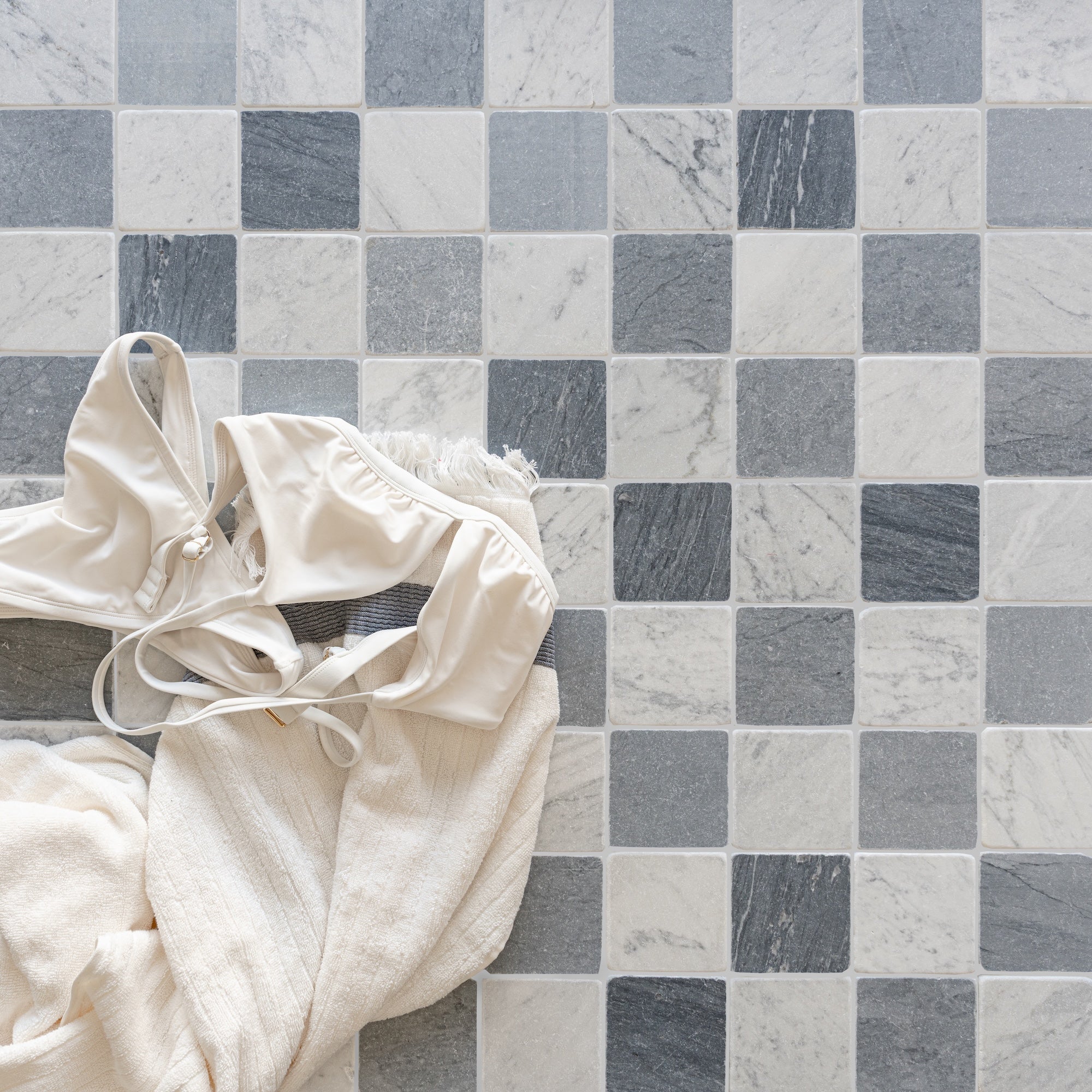 Square ceramic tiles in varying shades of gray and white forming a grid with cream fabric and swimwear draped on one side