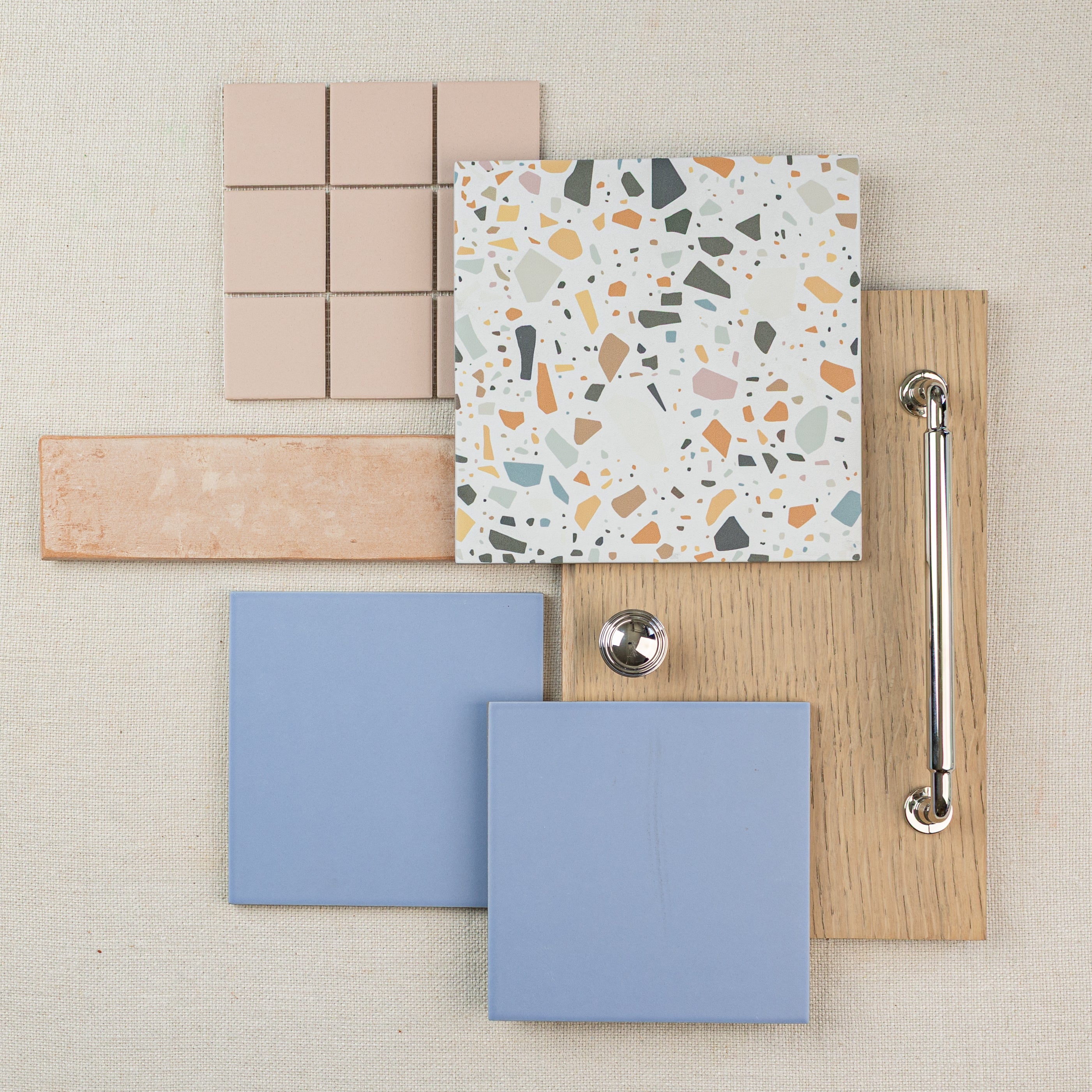 Flat lay of ceramic tile samples in beige, terrazzo, blue, and wood with a metal handle and knob on fabric background