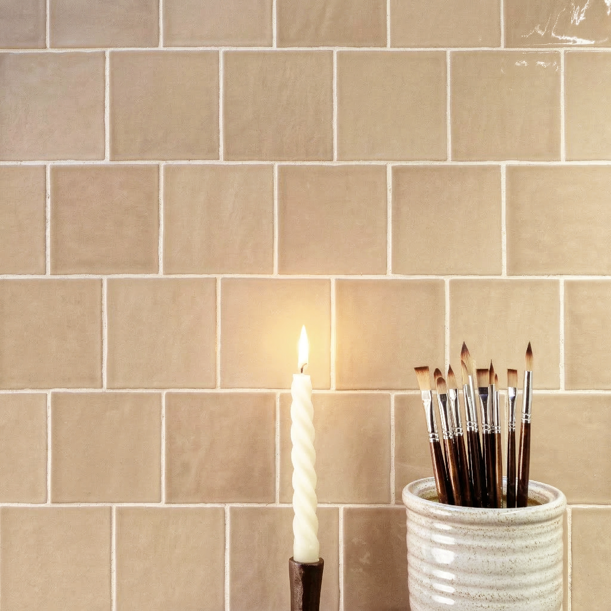 Glossy warm beige ceramic tiles arranged in a staggered pattern behind a lit twisted candle and ceramic brush holder