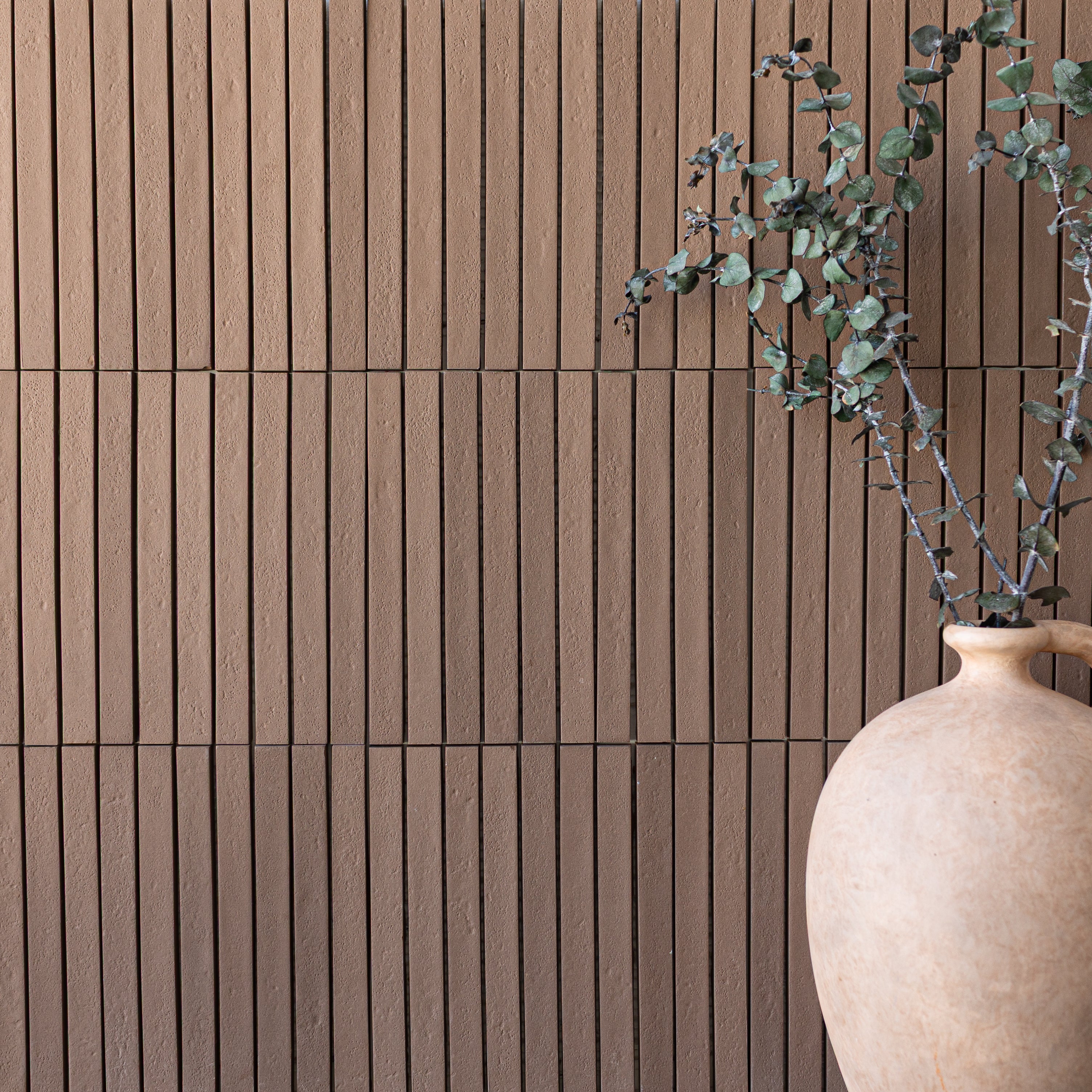 Vertical terracotta tiles with fine grooves create texture beside a matte beige ceramic vase holding eucalyptus branches