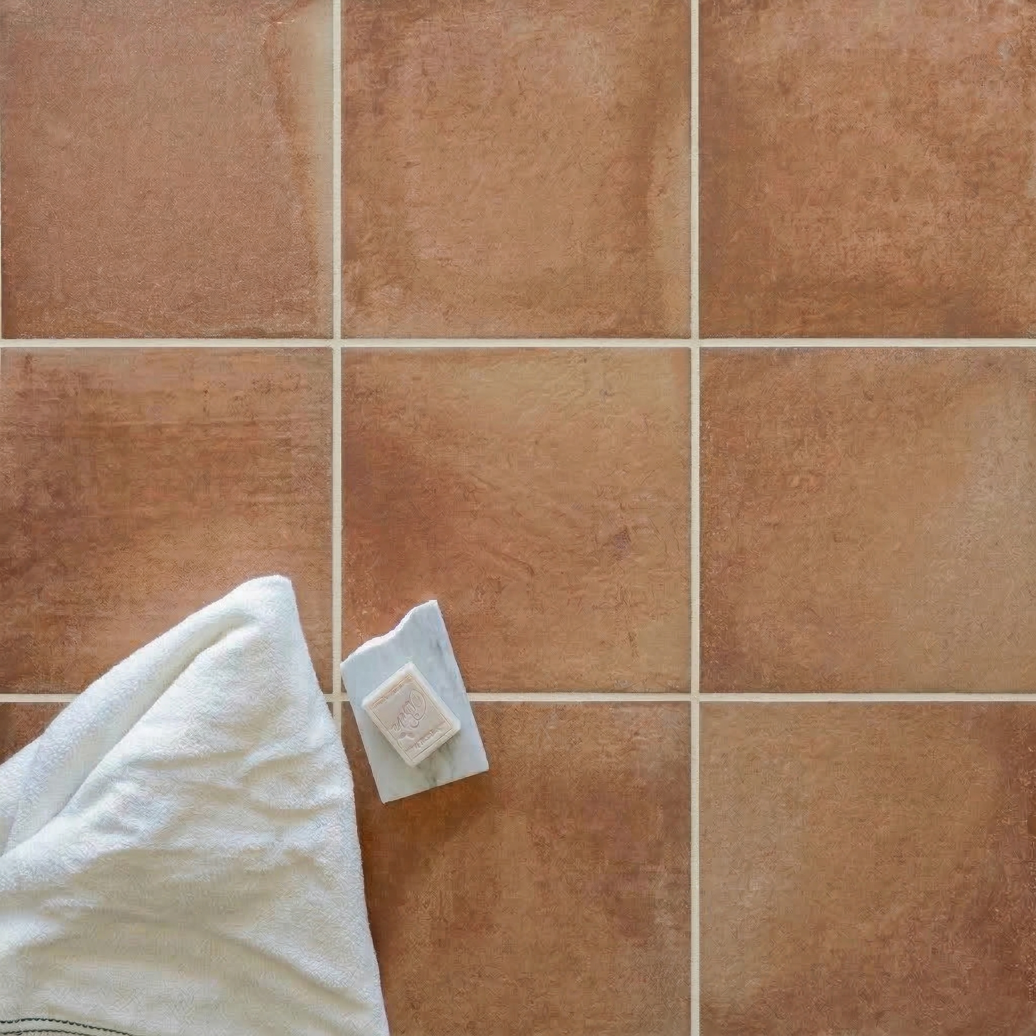 Rust-colored terracotta tiles with subtle texture arranged in a grid beside a folded white cloth and a small soap bar.