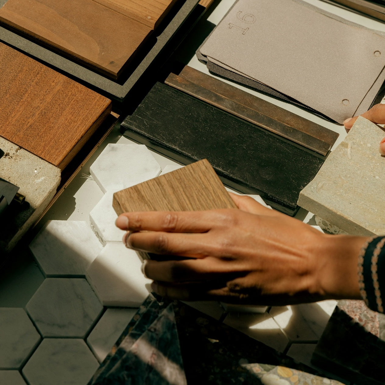 Hand holding wood veneer sample among hexagonal marble tiles, textured stones, and fabric swatches in natural light