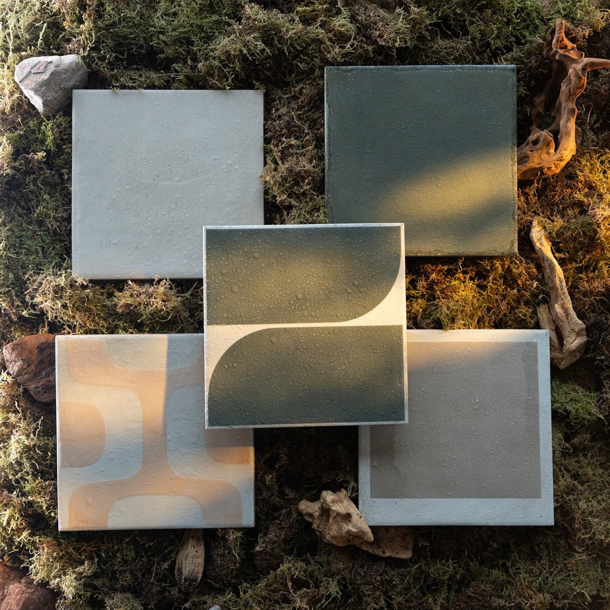 Five ceramic tiles in neutral tones with abstract designs arranged on a bed of natural moss and wood pieces