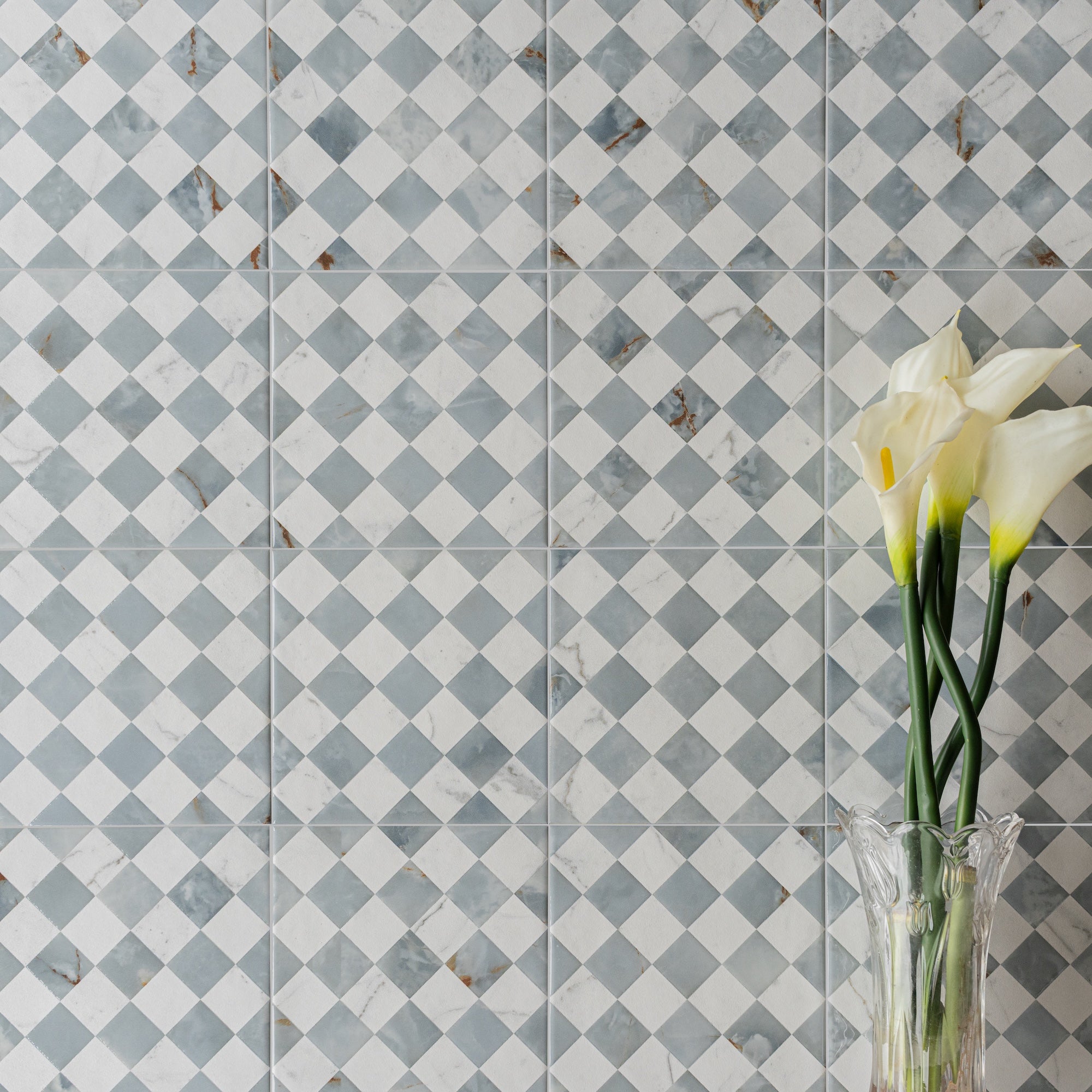 Subtle checkerboard ceramic tiles in soft gray and off-white with a clear vase holding delicate cream calla lilies