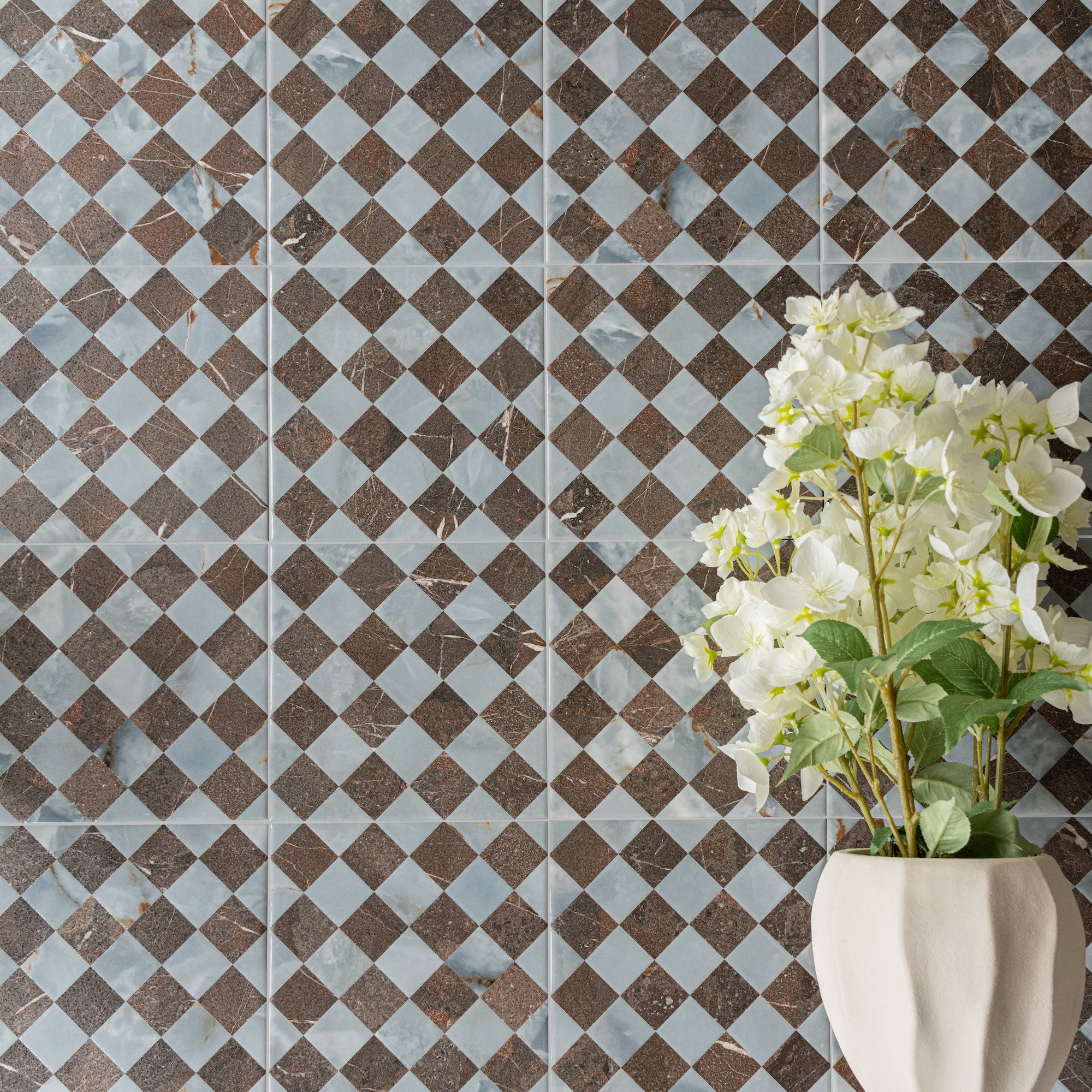 Checkerboard ceramic tiles in shades of brown and light blue with a white vase holding fresh white flowers against the wall