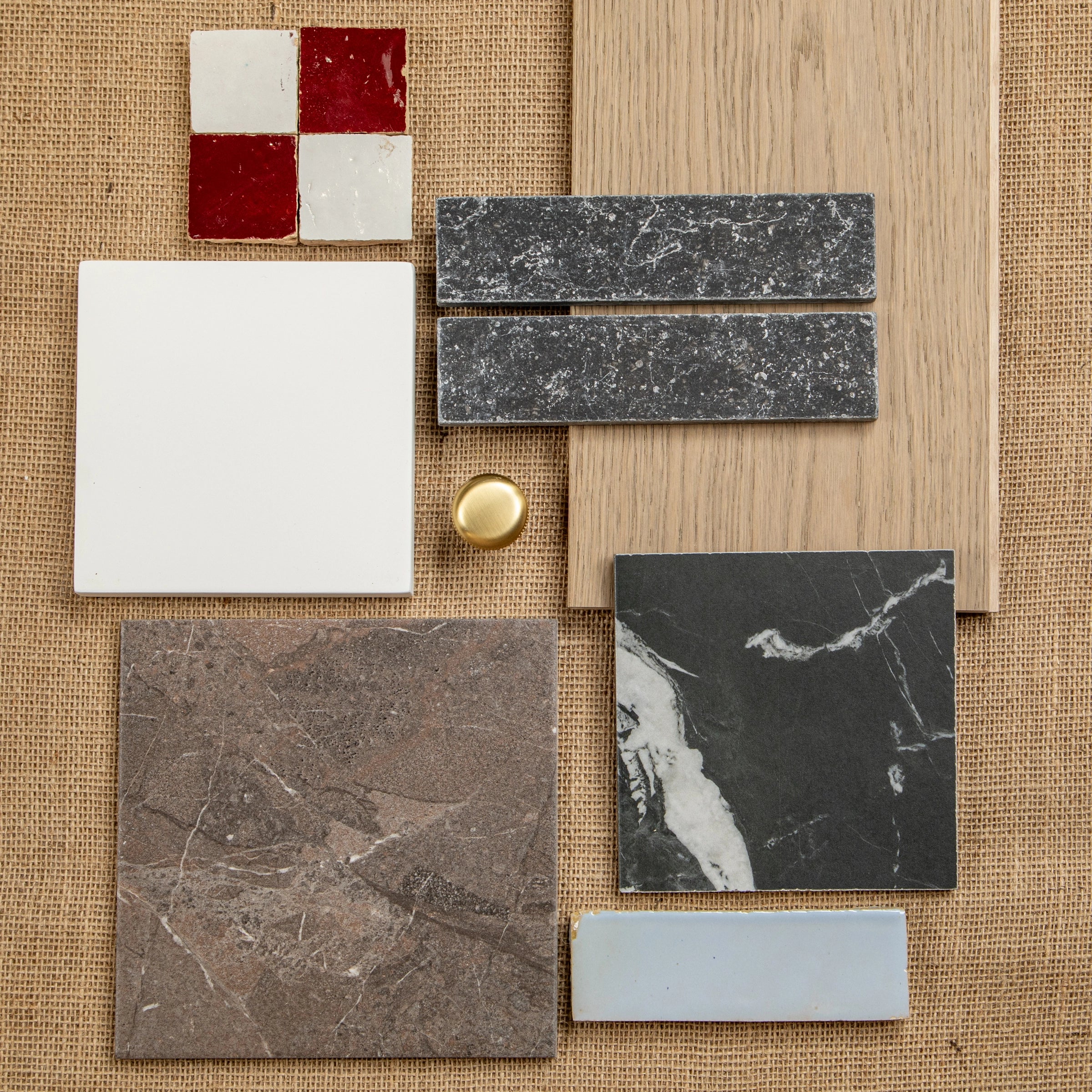 Flat lay of red and white ceramic tiles, black marble-look tiles, beige wood plank, gray stone samples, and brass knob on textured fabric