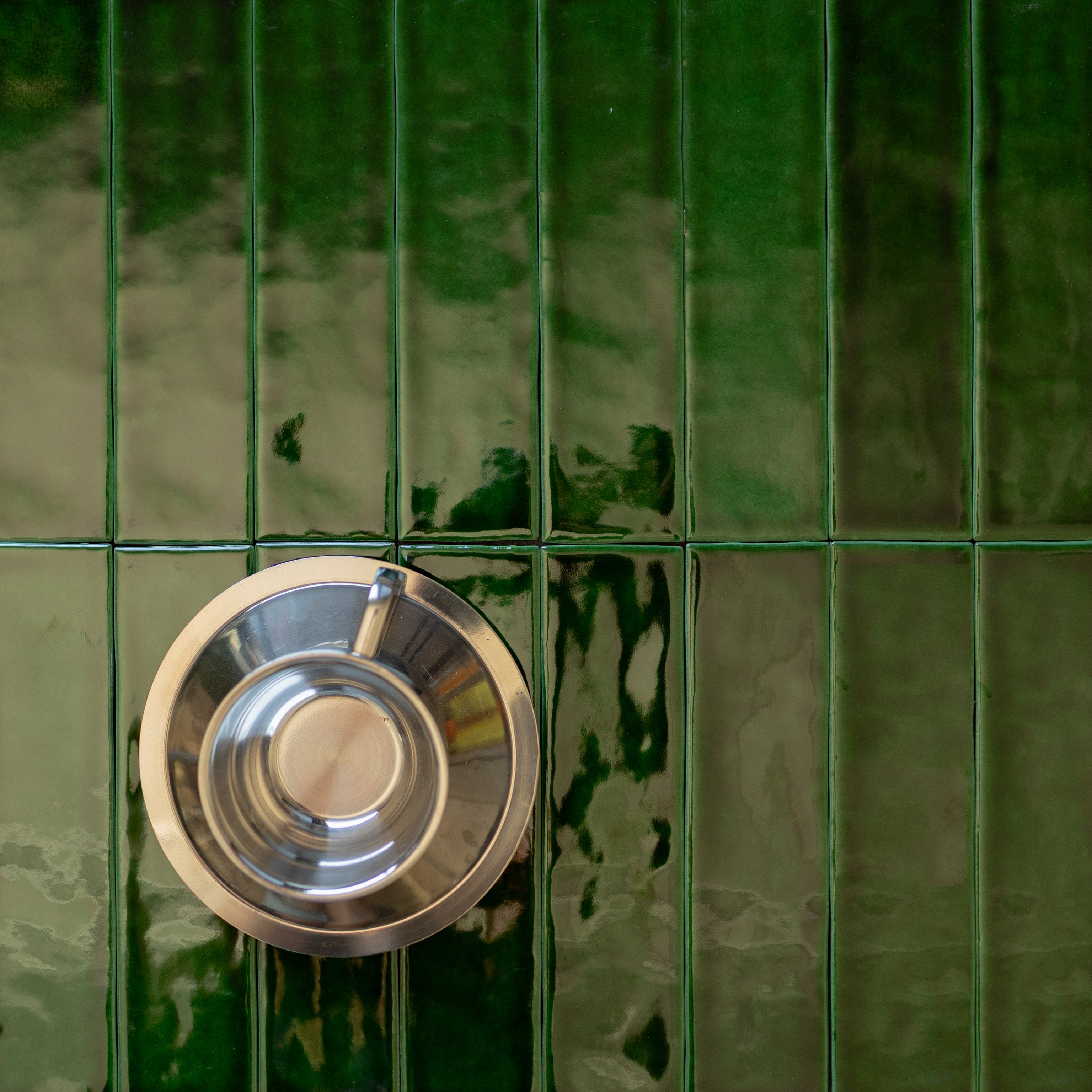 Glossy deep emerald ceramic tiles arranged vertically with a shiny silver metal cup resting on the surface