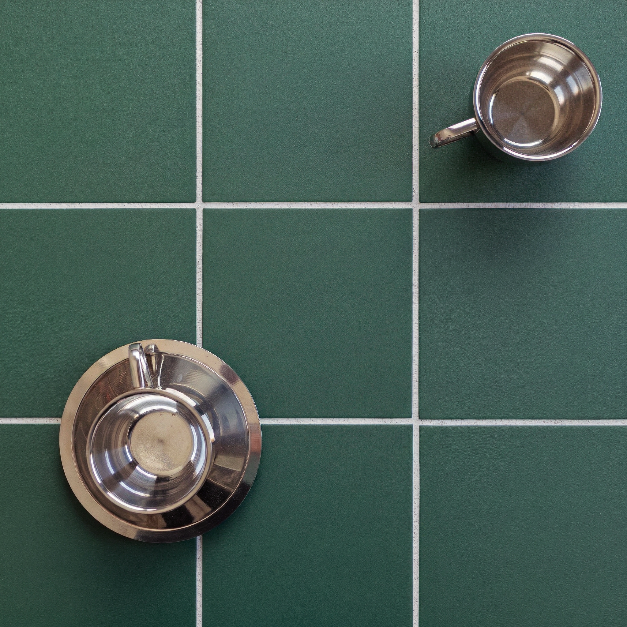 Top view of two stainless steel cups on green square ceramic tiles with white grout lines.