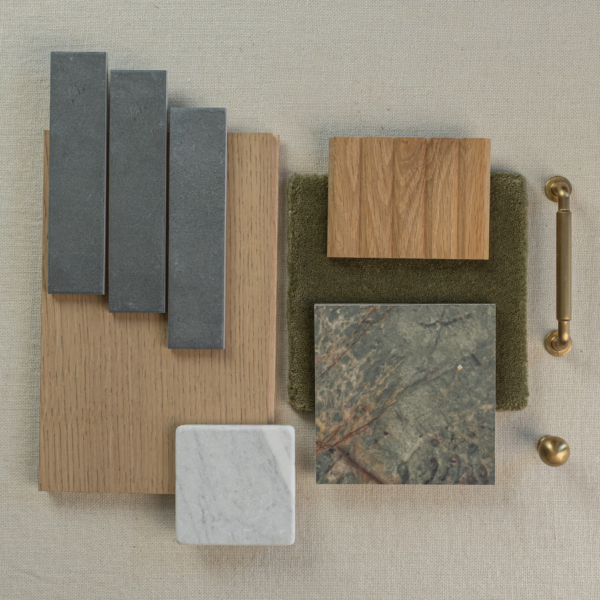 Flat lay of charcoal gray rectangular tiles, wood samples, textured green fabric, marble tile, and brass hardware on neutral fabric