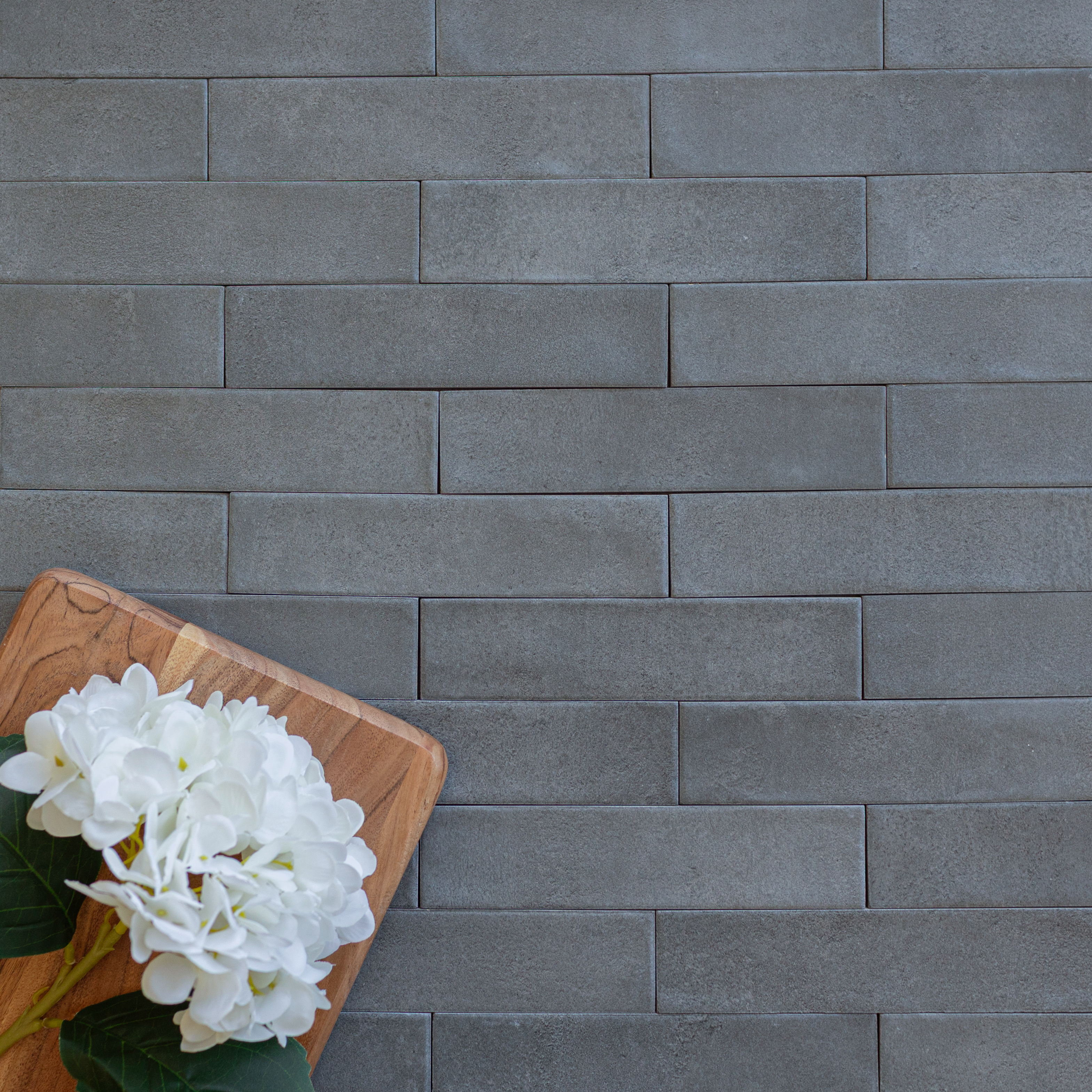 Matte charcoal rectangular ceramic tiles in staggered rows with a wooden board holding white flowers in the corner