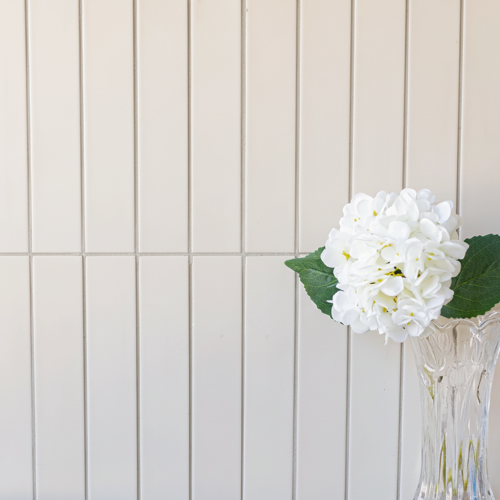 Glossy cream ceramic tiles arranged vertically paired with a clear vase holding a white hydrangea and green leaves