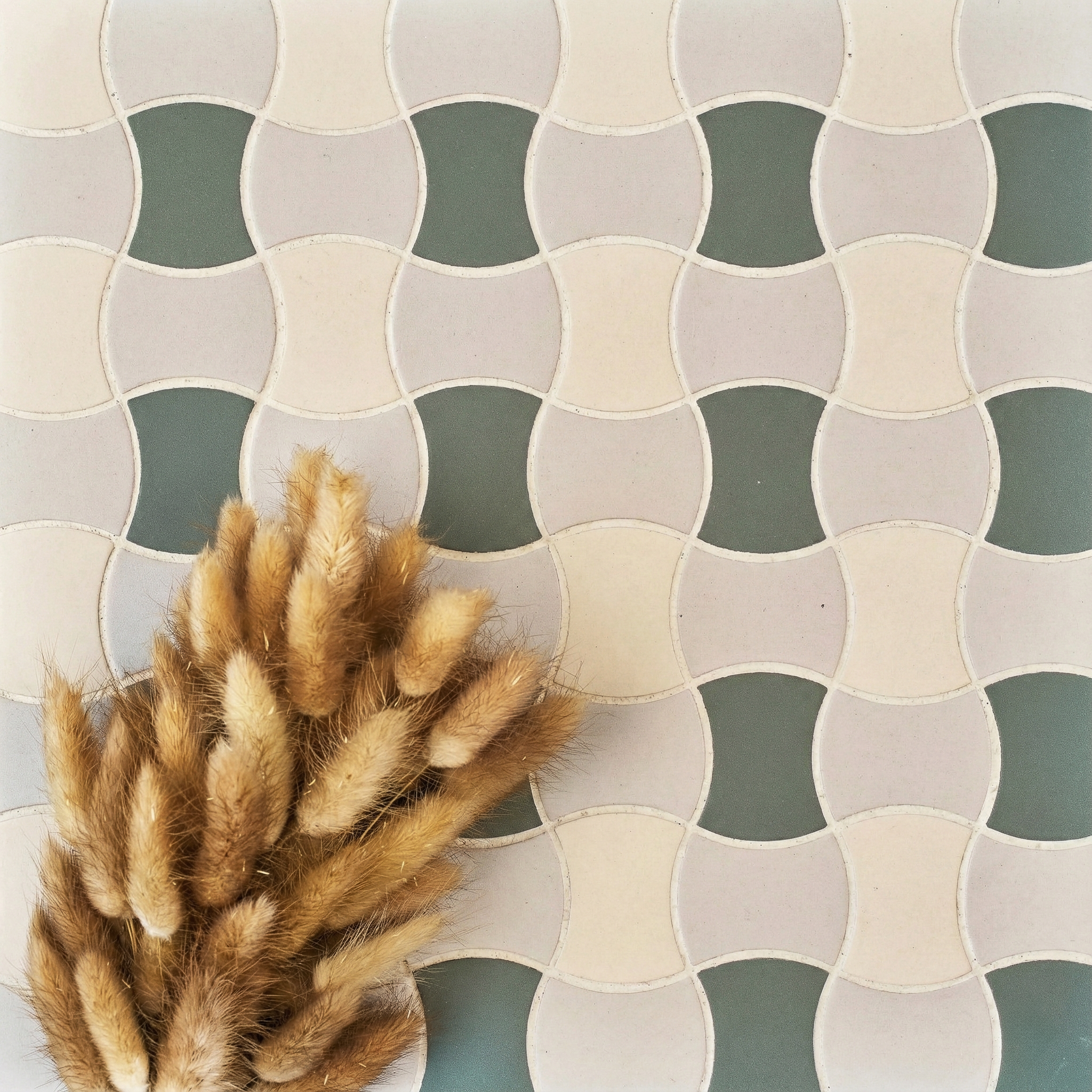 Curved ceramic tiles in soft green, cream, and beige tones arranged with dried wheat stalks on a pale surface