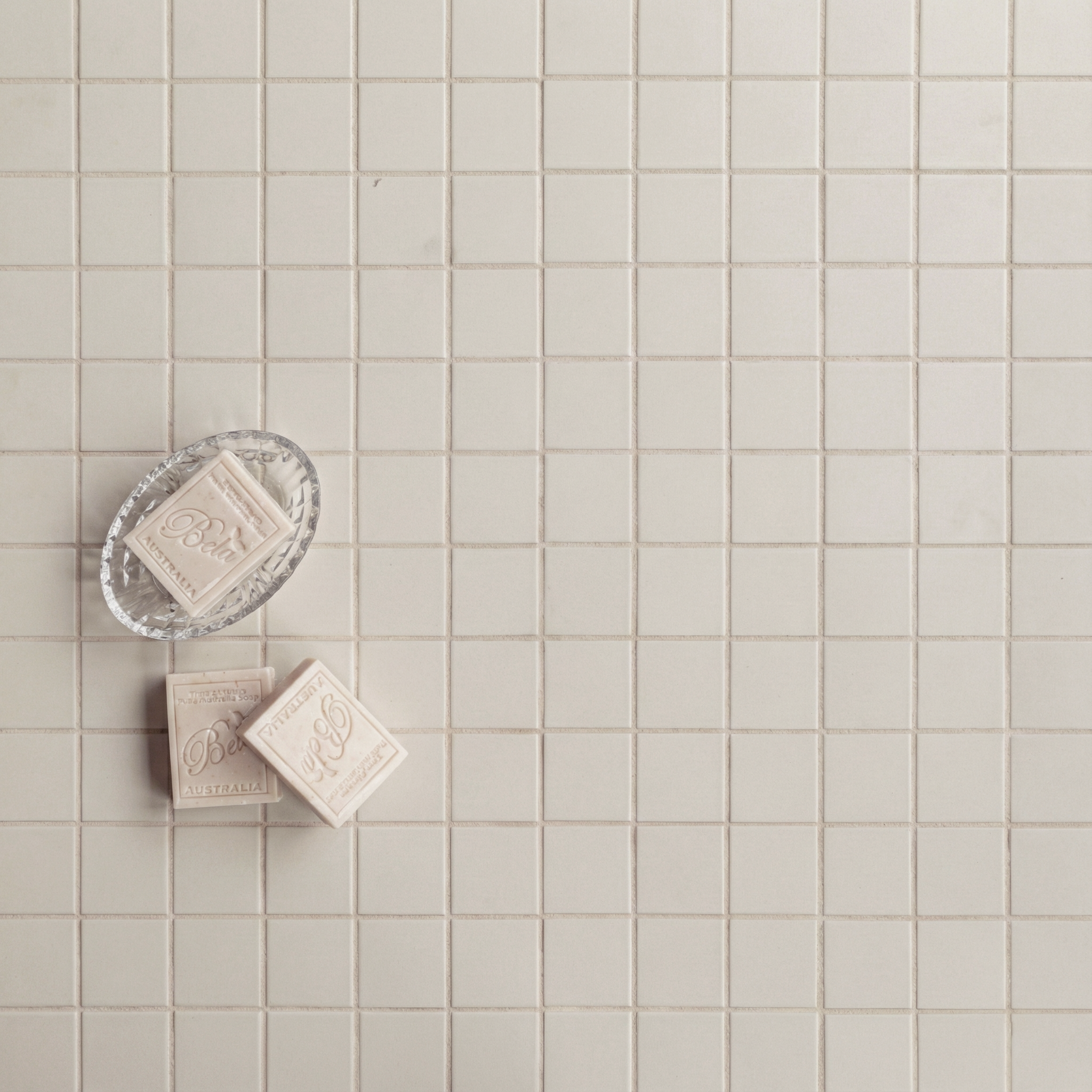 Grid of small matte cream ceramic tiles with three beige soap bars on a clear dish in the corner