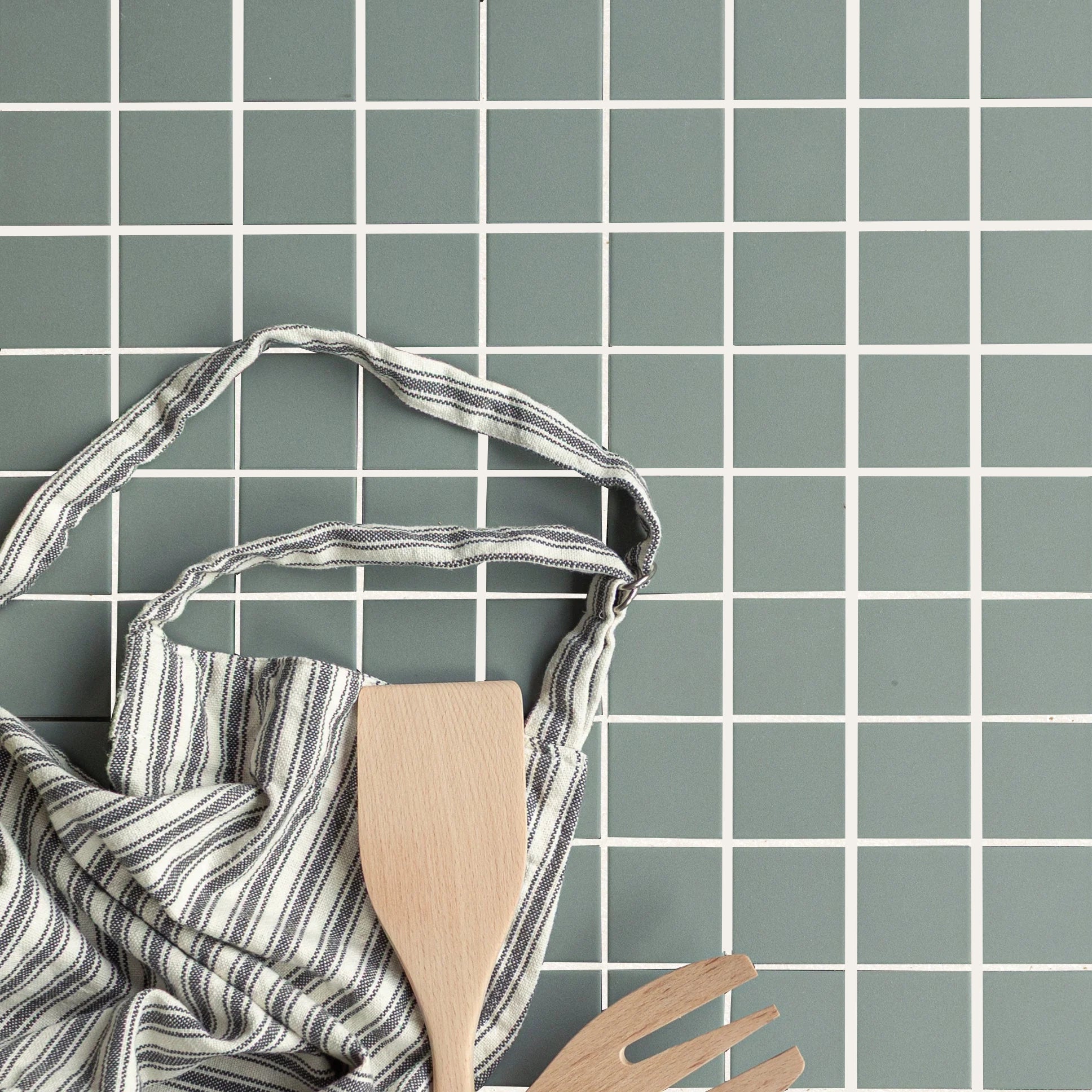 Matte muted green porcelain mosaic tiles with light grout lines and striped kitchen towel with wooden utensils resting on surface