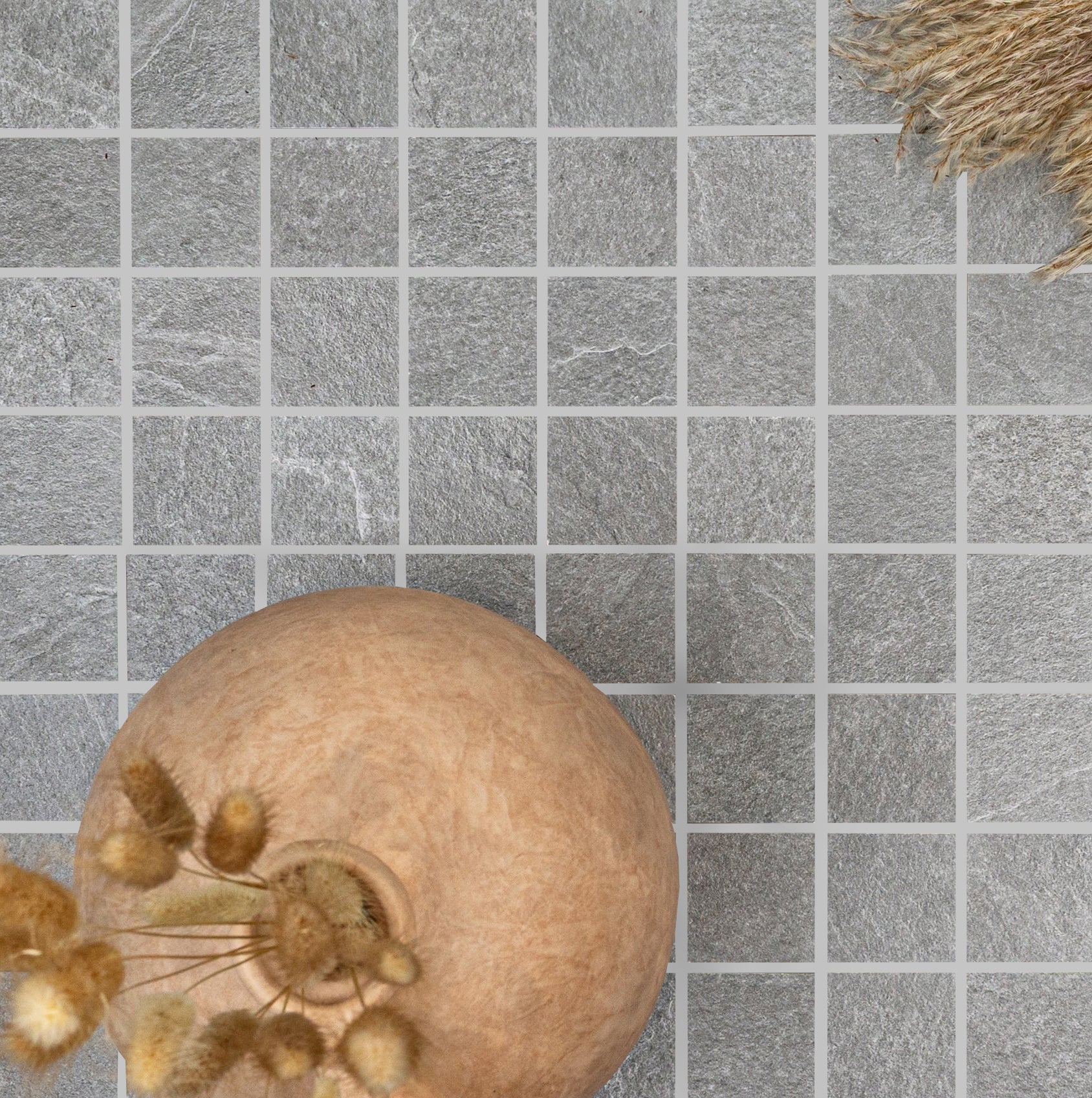 Close-up of textured gray slate tiles arranged in a grid pattern with a round clay vase and dried grasses nearby