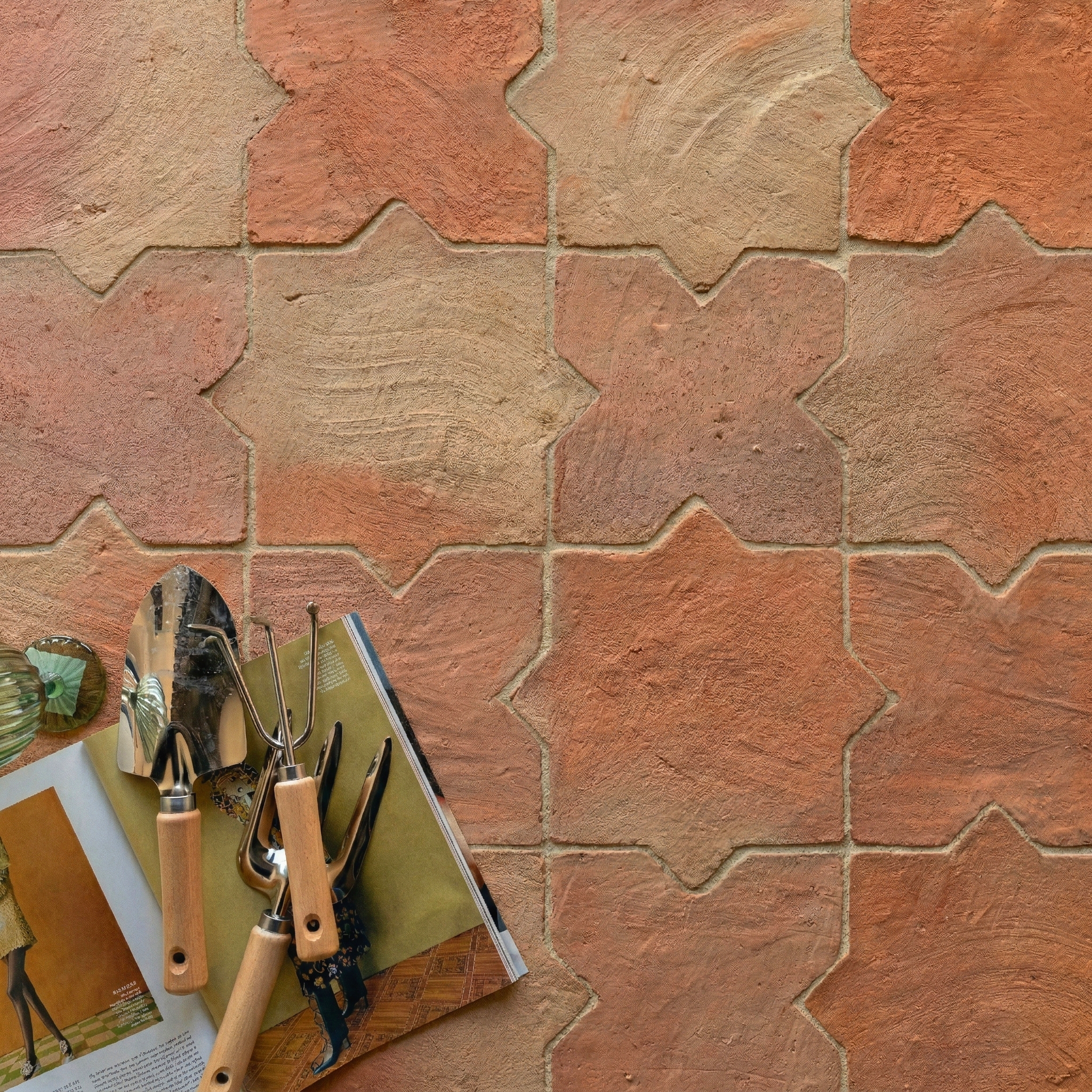 Rustic terracotta tiles with star and cross shapes in warm earth tones next to small gardening tools and a magazine.