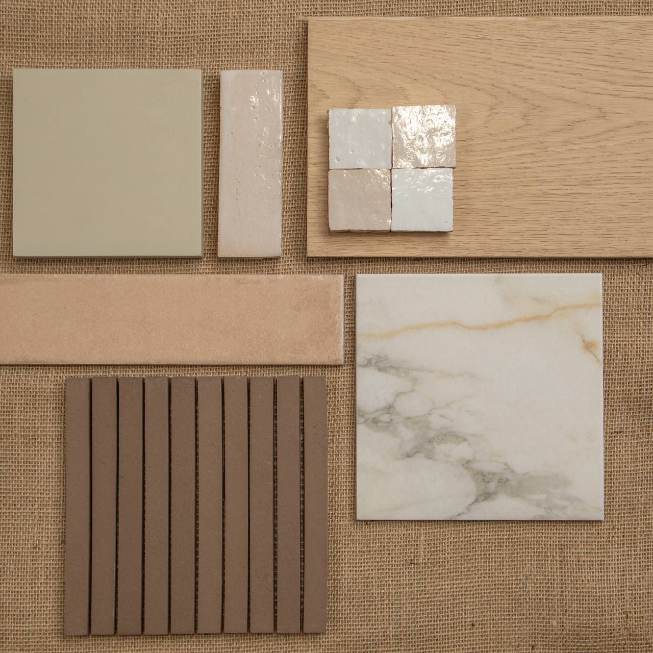Collection of beige and cream ceramic tiles, wood plank sample, and textured brown tile arranged on natural fabric background