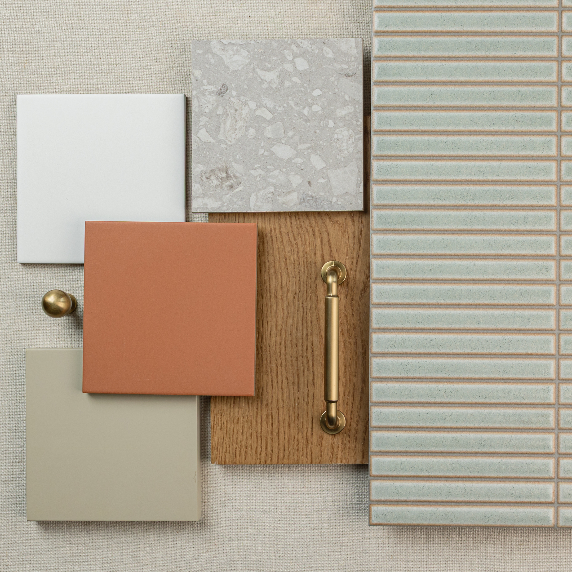 Flat lay of ceramic tiles, wood sample with brass handle, linear ceramic mosaic tile, and brass knob on neutral fabric background