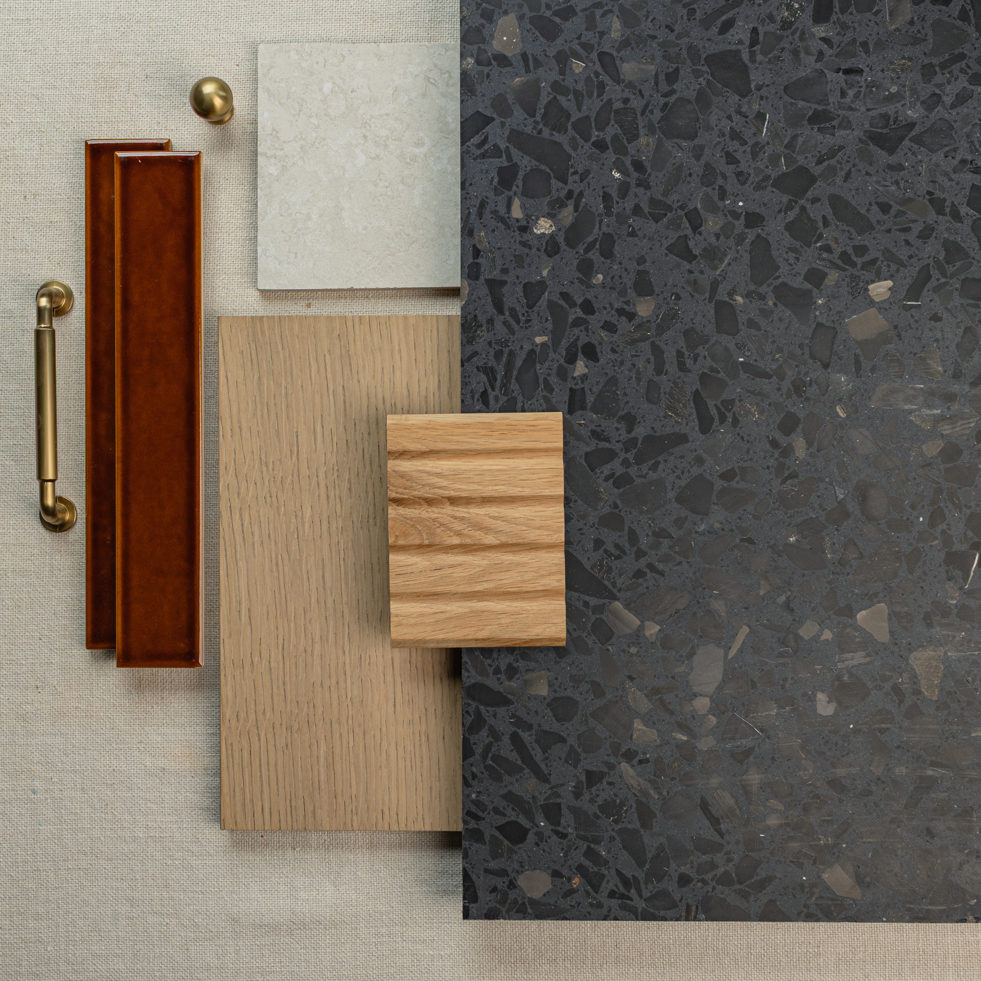 Dark terrazzo tile, light oak wood samples, a beige tile, and brass handles on neutral textured fabric background