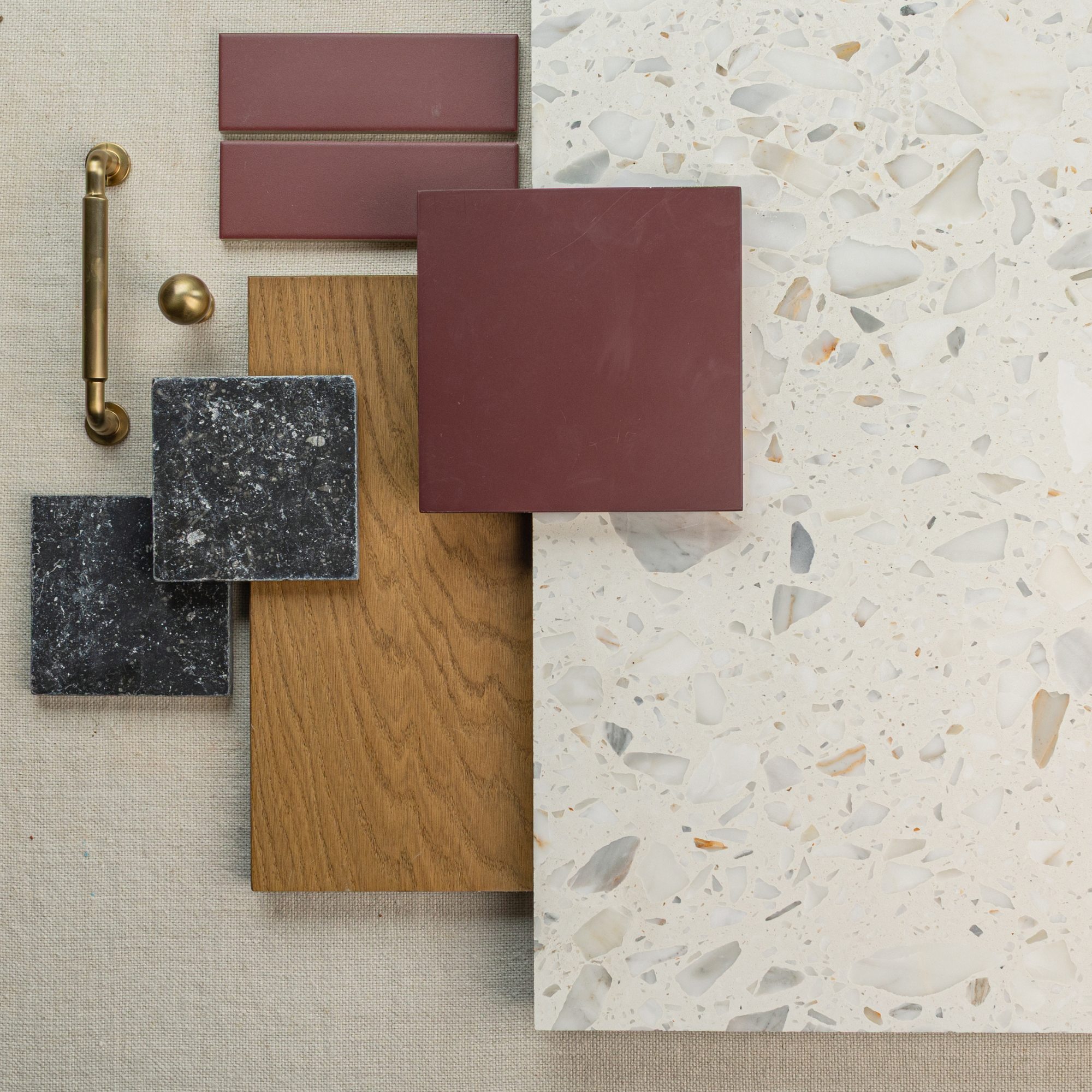 Arrangement of matte red ceramic tiles, dark speckled stones, wood plank, brass hardware, and a terrazzo surface panel