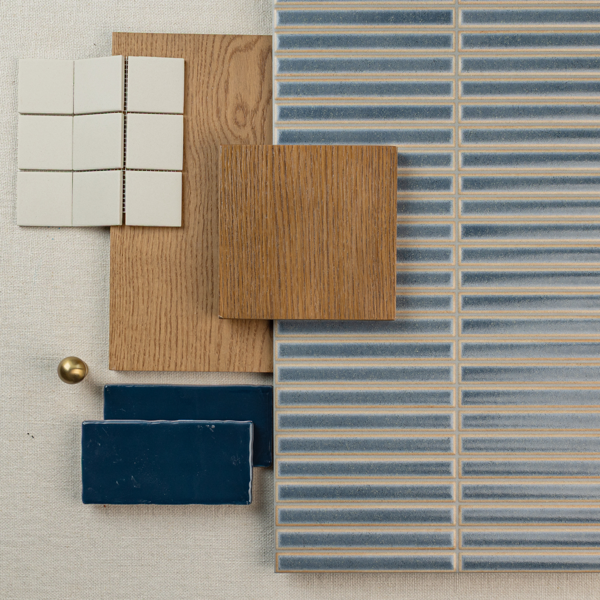 Flat lay featuring blue striped ceramic tiles, natural oak wood samples, white square tiles, navy tile, and brass knob on neutral fabric background