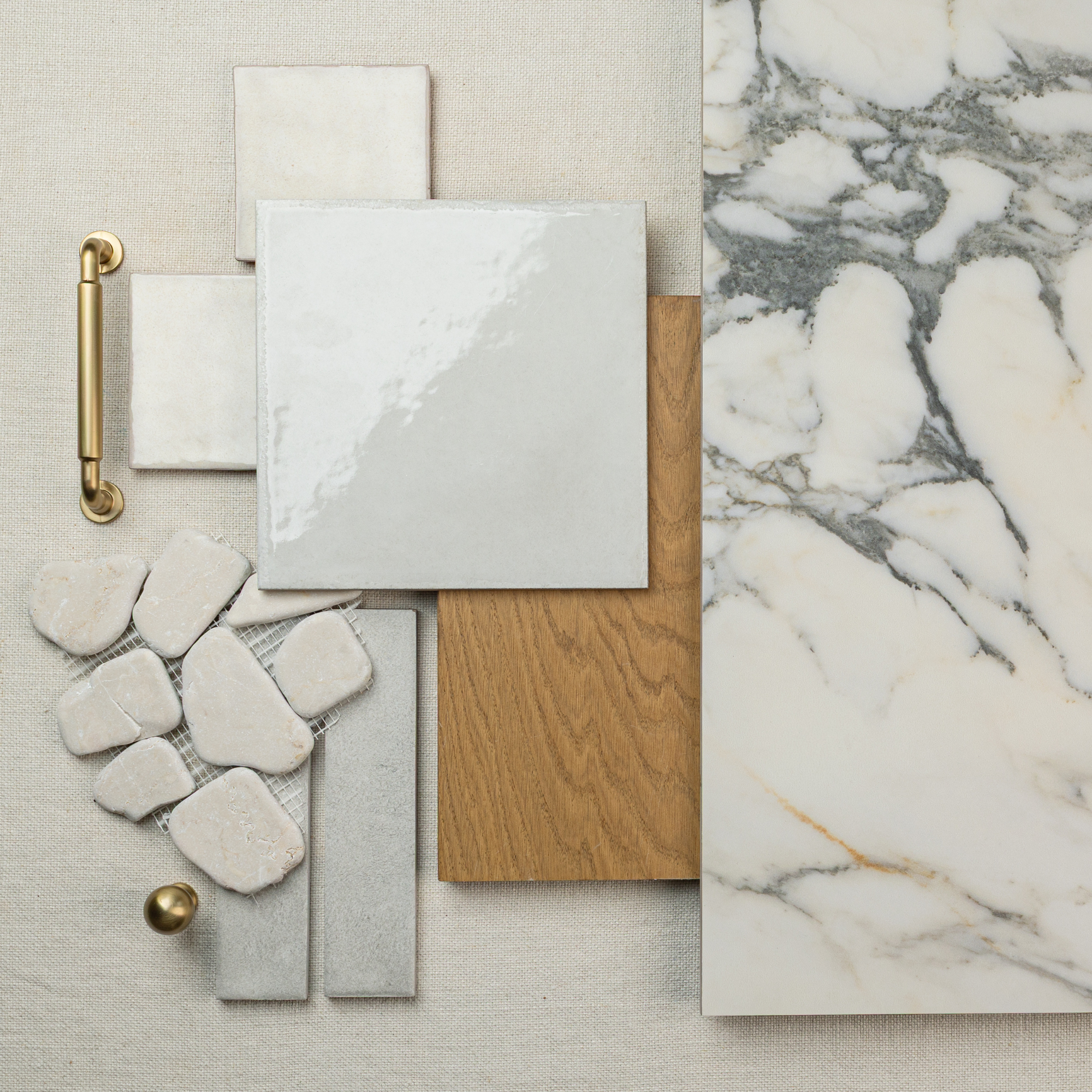 Arrangement of large marble-look tile, wood plank sample, cream ceramic tile samples, stone mosaic, and brass hardware on neutral fabric